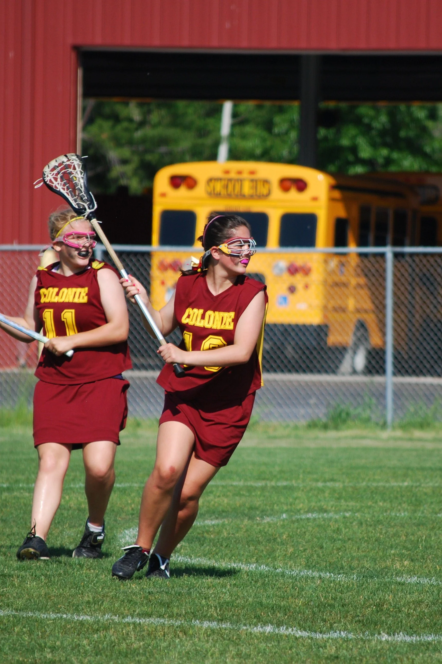 Two young female lacrosse players in maroon and yellow uniforms playing on a field, with a yellow school bus and a red building in the background.