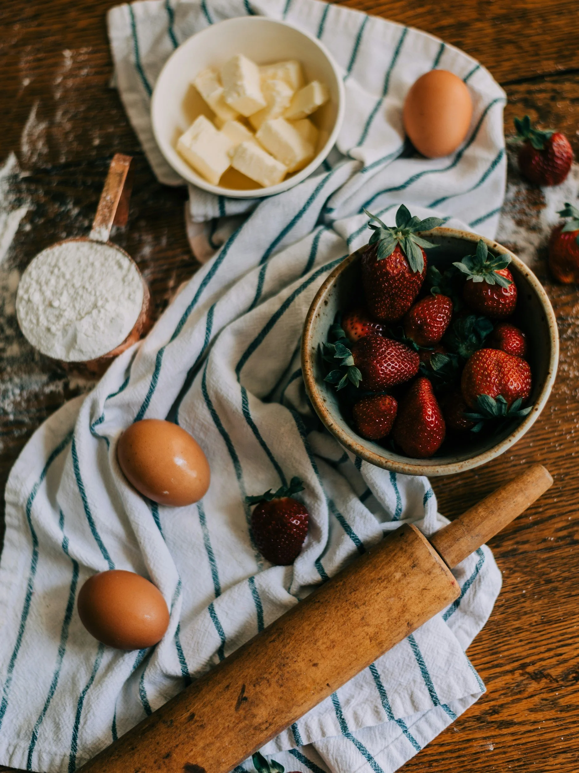 Fresh strawberries, eggs, butter, flour, and a rolling pin on a striped cloth on a wooden surface.