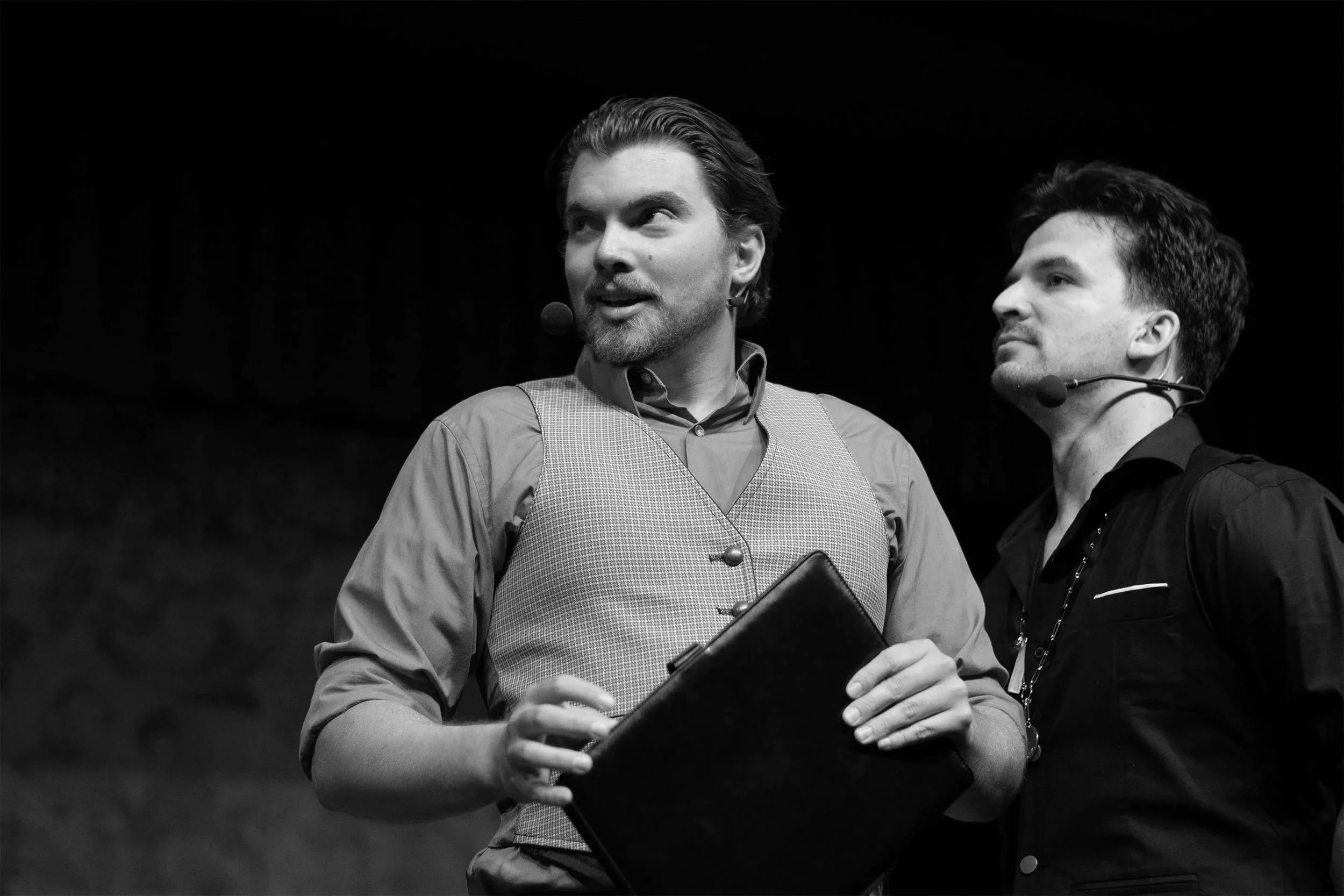 Two men with headsets speaking on stage, one holding a clipboard, against dark background. Black and white photo.