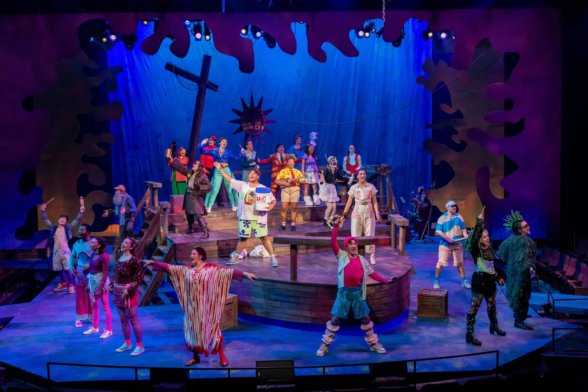 A colorful stage musical production with actors in vibrant costumes performing a lively scene with set pieces like a wooden cross and a sign that reads 'The Baton End'.