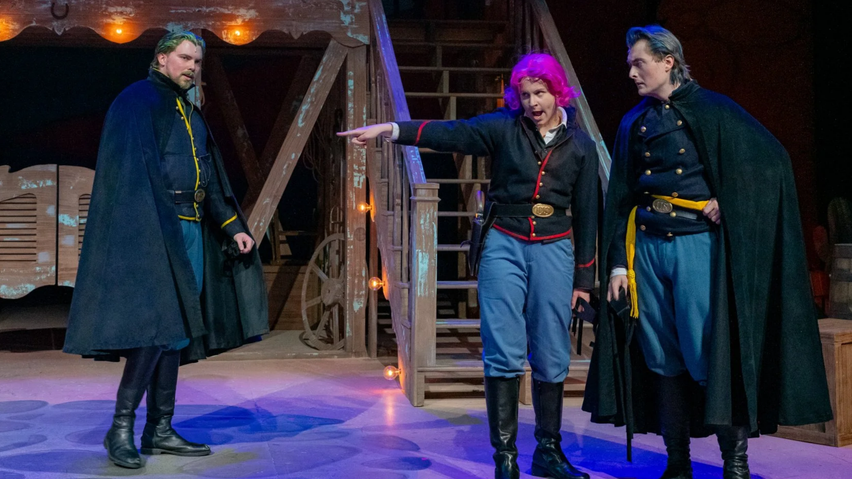 Three actors on stage performing in a play, with a wooden set resembling a rustic house and stairs in the background. The woman in the middle, with bright pink hair, is pointing and appears to be speaking. The men on either side wear dark capes and military-style uniforms, with one having gray hair and the other a beard.