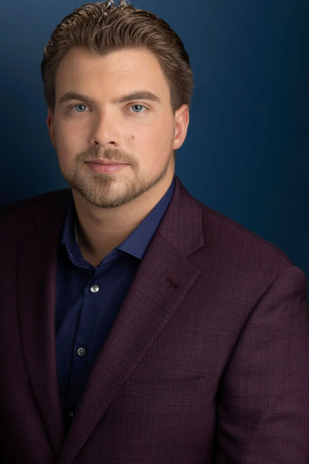 Headshot of a man with blond hair and blue eyes, wearing a dark blue shirt and a maroon blazer, against a dark blue background.