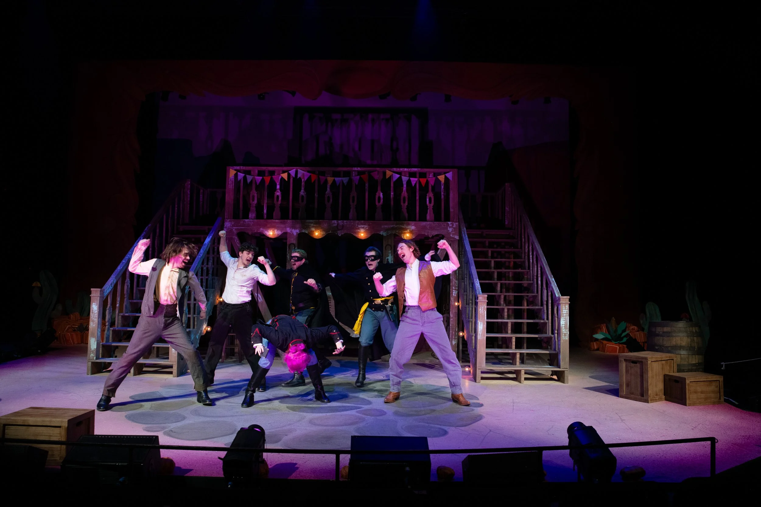 Theater stage with cast performing a lively scene, stairs in the background decorated with colorful bunting, stage lighting, and props scattered around.