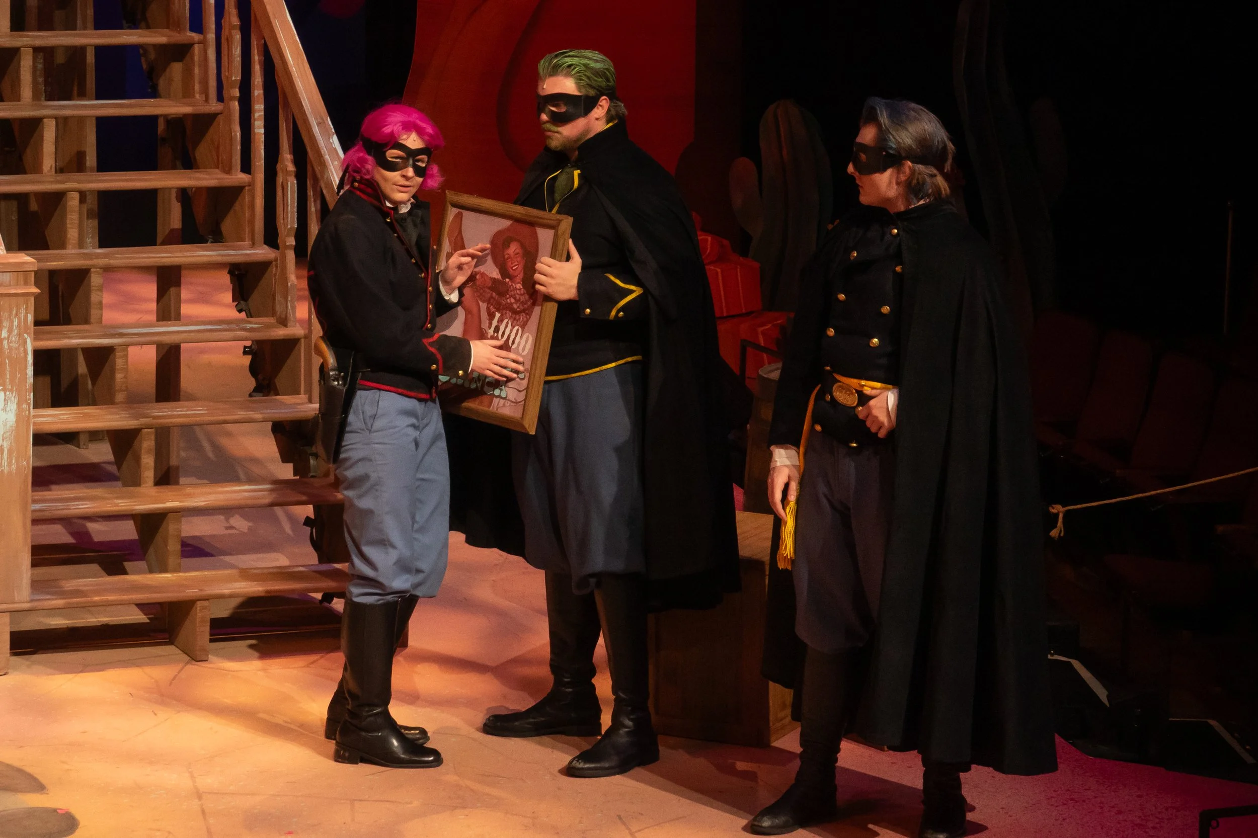 Three actors dressed as masked bandits in costume, with one holding a framed vintage portrait, standing on stage near wooden stairs and red velvet furniture.