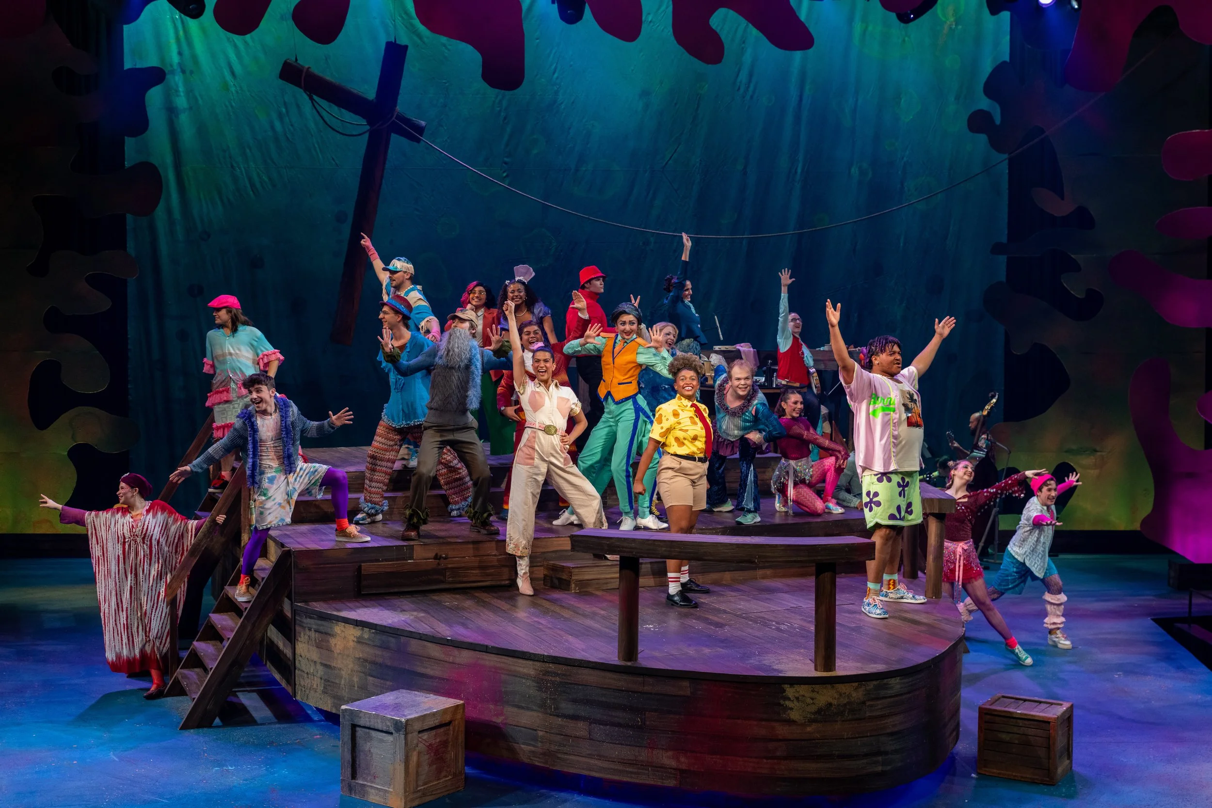 A lively stage scene from a theatrical musical with colorful costumes, featuring a diverse group of performers dancing and singing on a wooden ship set.