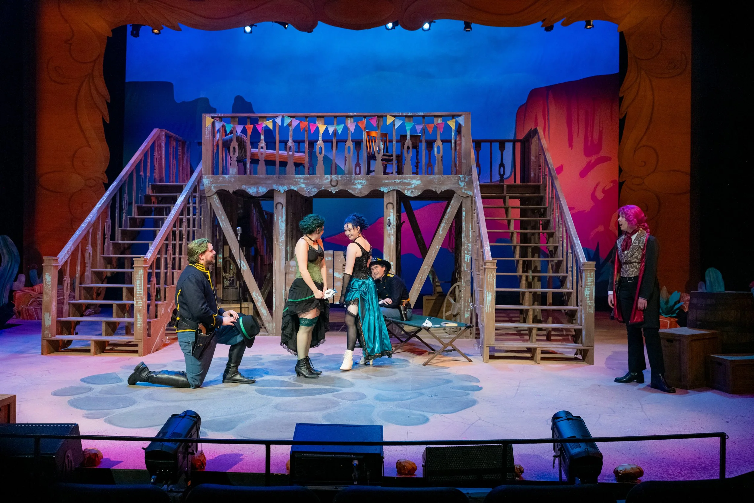 Theater stage set with wooden platform and stairs, colorful bunting, and painted backdrop. Cast members are in costume, with some kneeling and some standing, engaging in a scene.