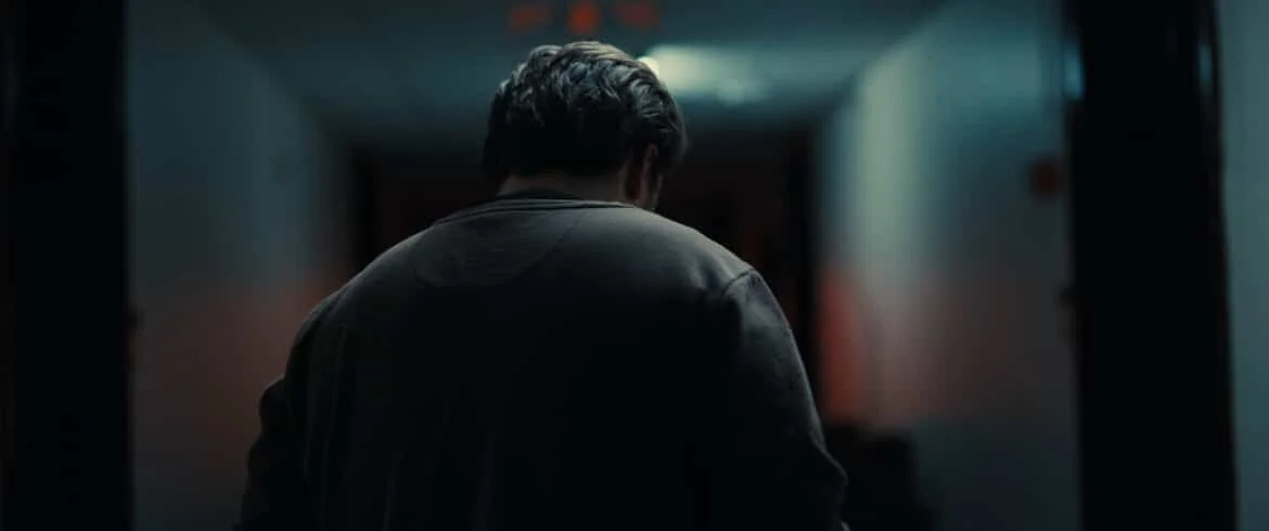 A person with dark hair, seen from behind, walking down a dimly lit hallway.