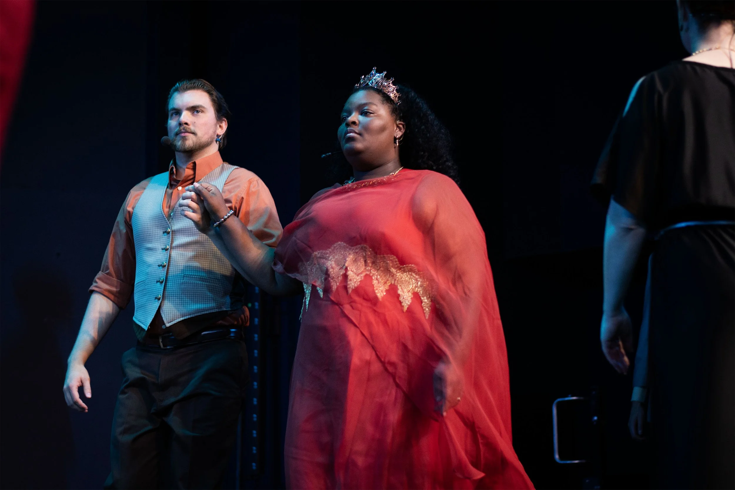 A man and a woman standing close together on a stage, possibly during a performance or ceremony. The man has light skin, dark hair, and a beard, and is wearing a tan and blue vest with a long-sleeved shirt. The woman has darker skin, curly black hair