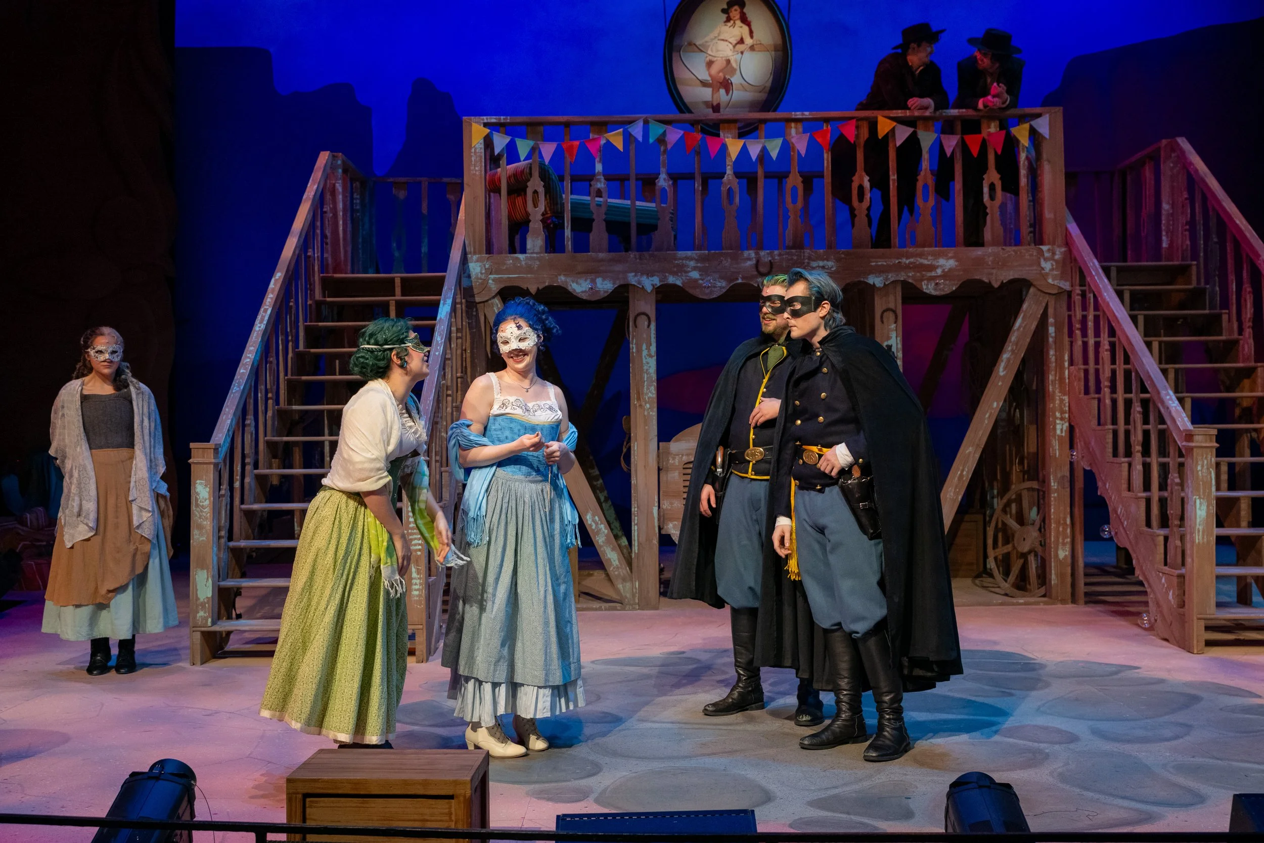 Actors in a theater production dressed in period costumes, with some wearing masks, on a set featuring a wooden staircase and balcony, with a painted portrait of a woman in the background.