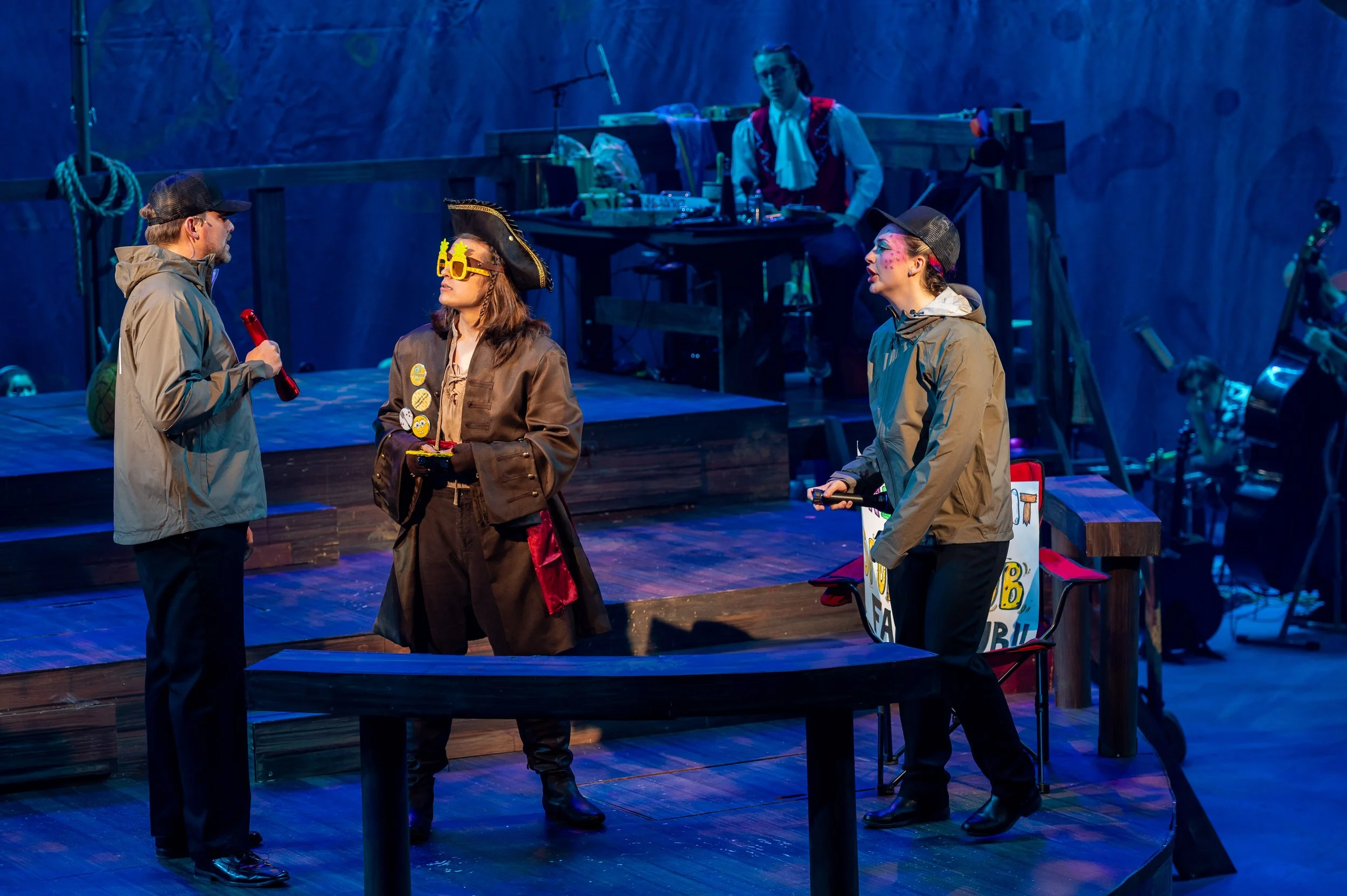 People in costumes performing on a stage with blue lighting, including a person dressed as a pirate, two people in casual jackets, and a person in formal pirate attire with glasses and badges, while a musician is visible in the background.