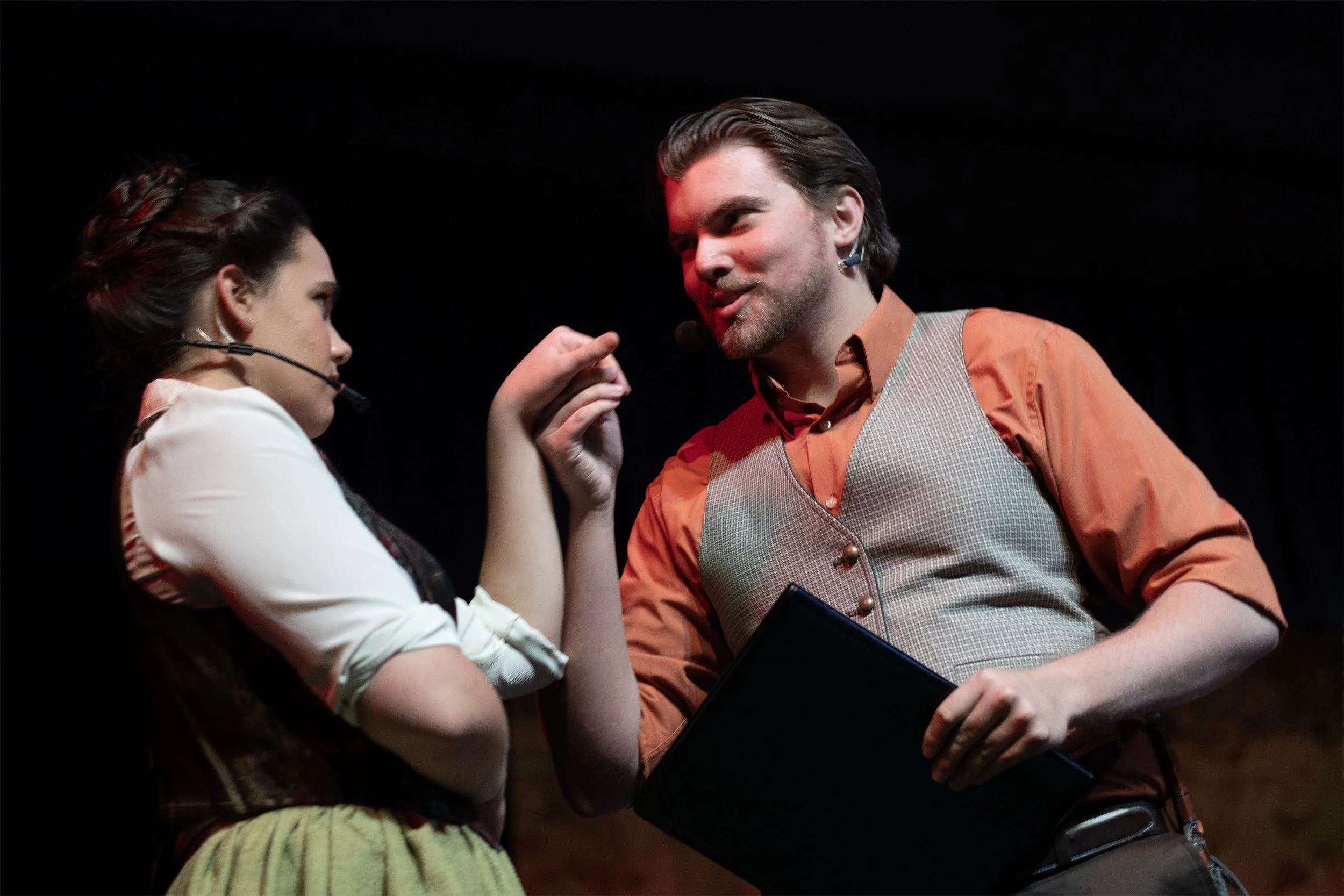 A man and a woman on stage engaged in a theatrical performance. The woman is wearing a white blouse with rolled-up sleeves, a dark vest, and a yellow skirt. She has dark hair styled in an elegant updo and is wearing a headset microphone. The man is dressed in an orange shirt with rolled-up sleeves, a checkered vest, and brown pants, holding a black folder. He has shoulder-length hair, a beard, and a headset microphone. They are making animated gestures and expressions against a dark background.