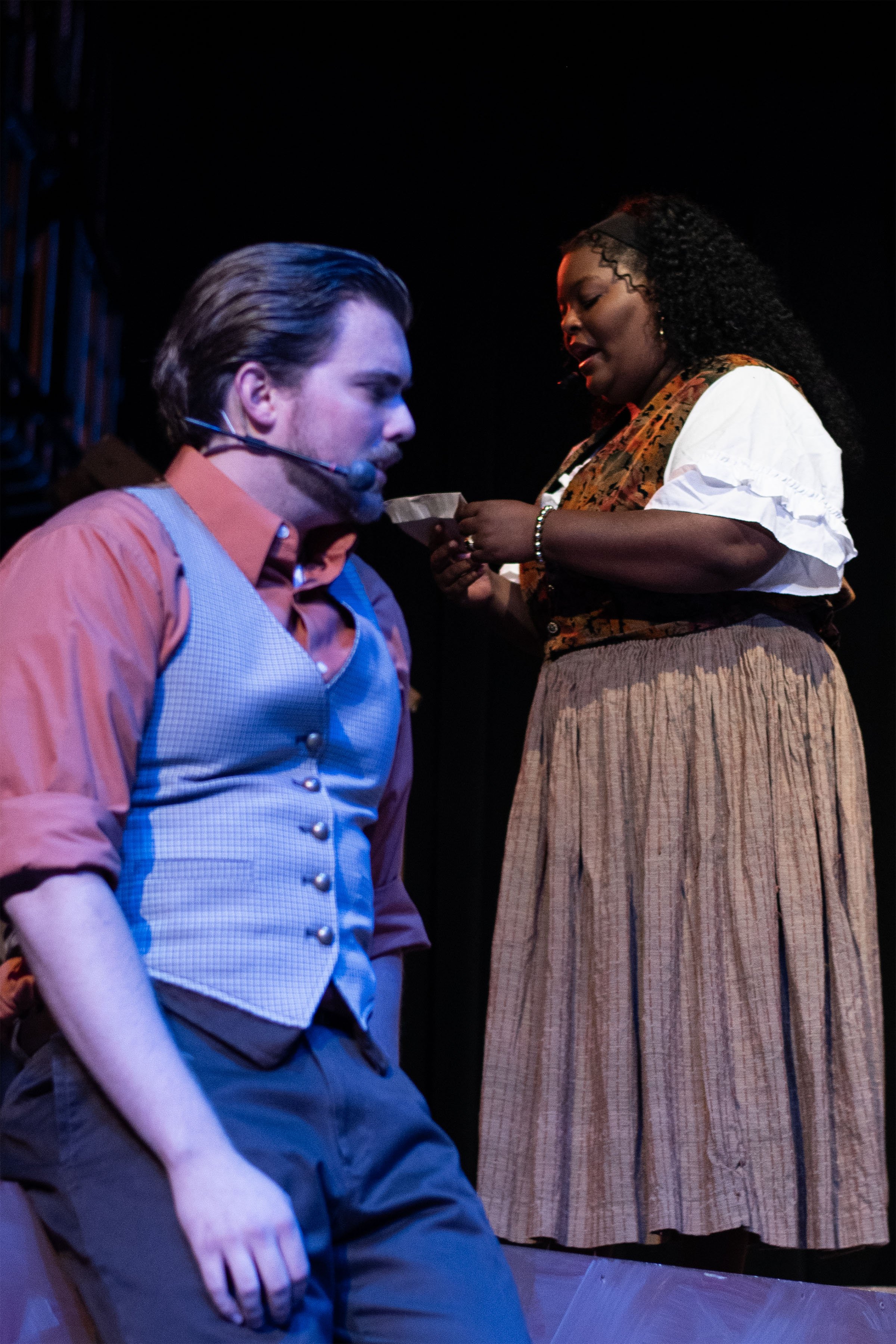 A man wearing a vintage vest and orange shirt kneels while a woman dressed in historical clothing stands, holding a small object in her hands during a theatrical performance.