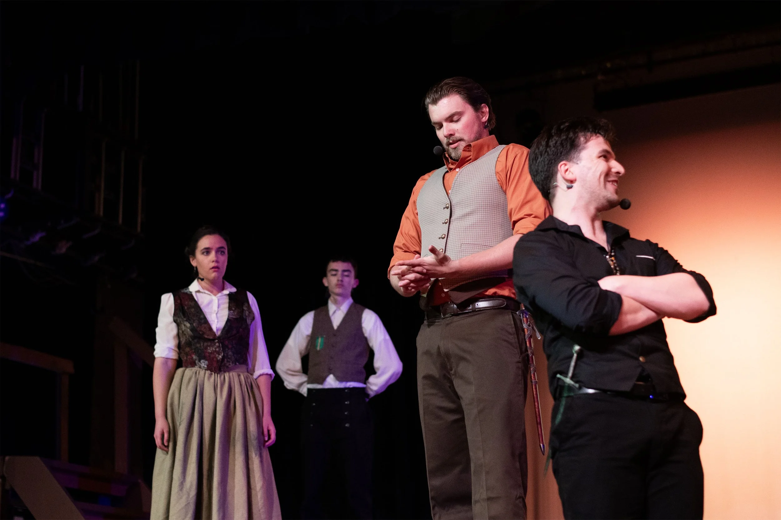 A group of four young actors on stage during a theatrical play. One woman in period costume, two young men in modern attire, and another woman in period costume, are visible while the man in modern attire is sitting and smiling.