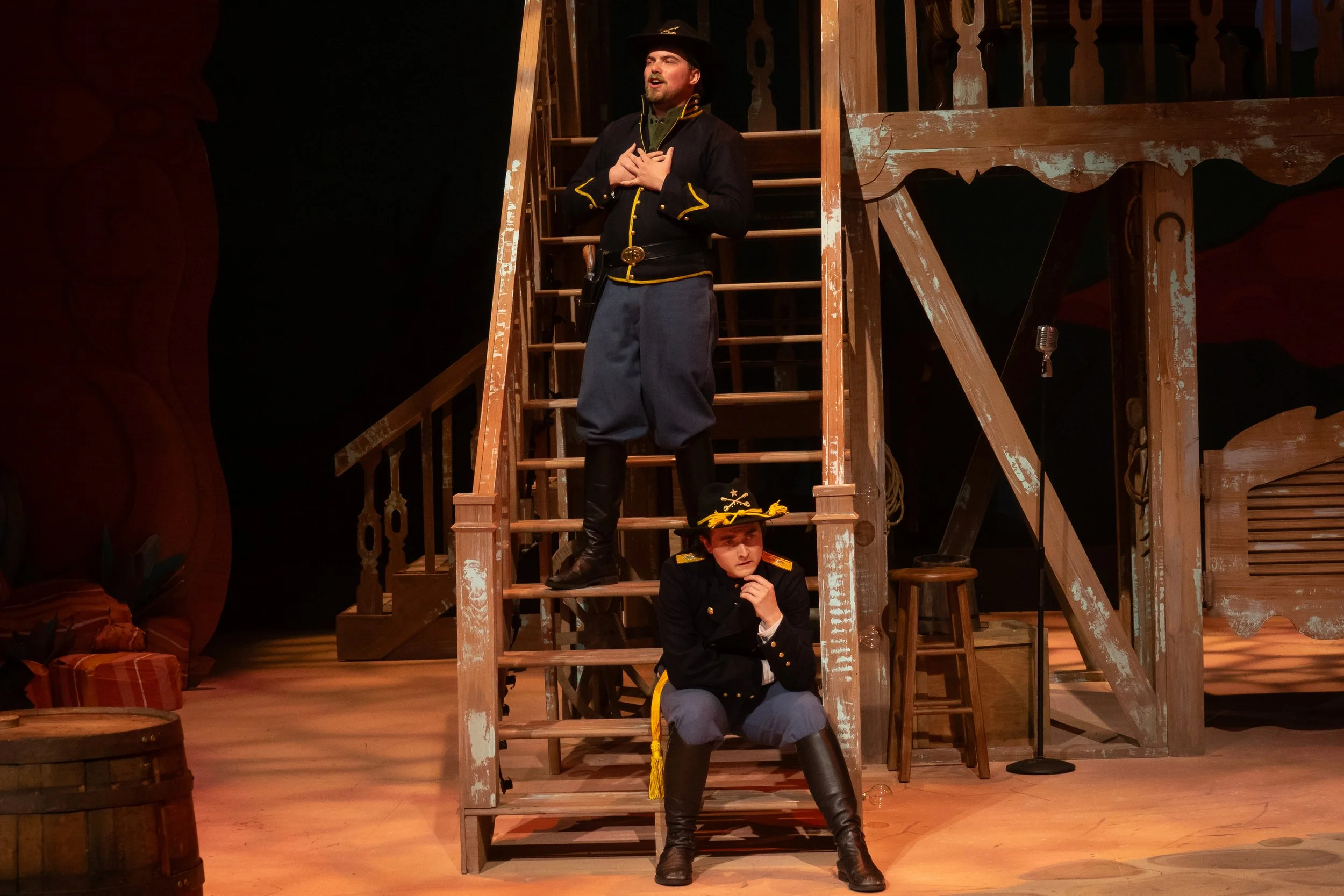 Three performers dressed in old-fashioned military costumes on a theater stage with wooden stairs and props, engaged in a scene with two standing and one sitting.