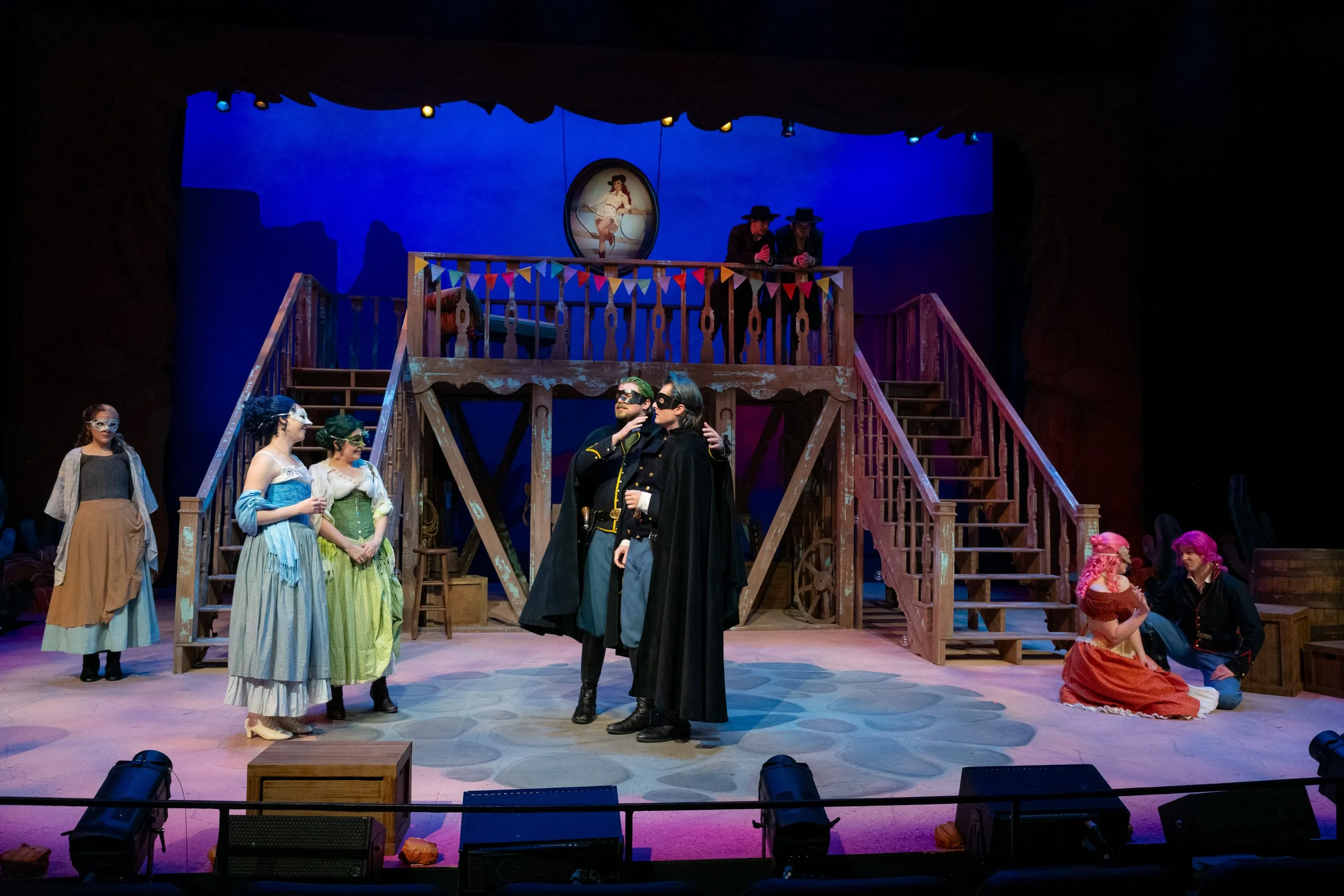 Theater stage with actors in period costumes, including women in dresses and men in military-style uniforms, performing a scene with a wooden balcony and colorful bunting, under theatrical lighting.