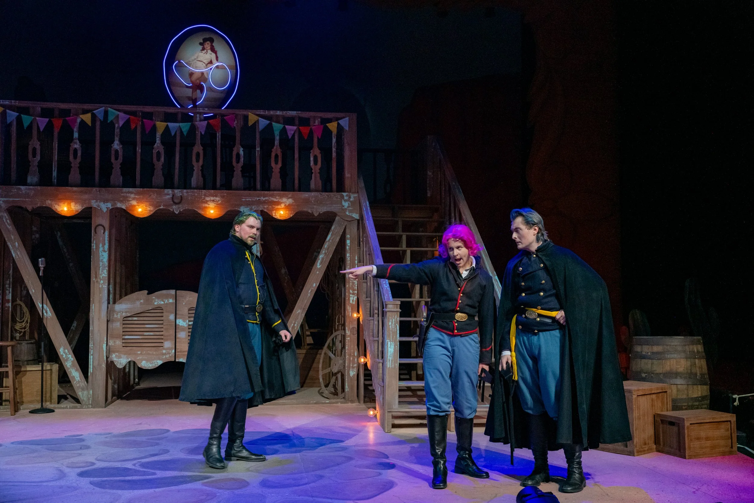Three actors in period costumes on stage, one woman with pink hair and two men with dark coats, in a theatrical setting with wooden stage structure and a neon sign of a woman in vintage attire in the background.