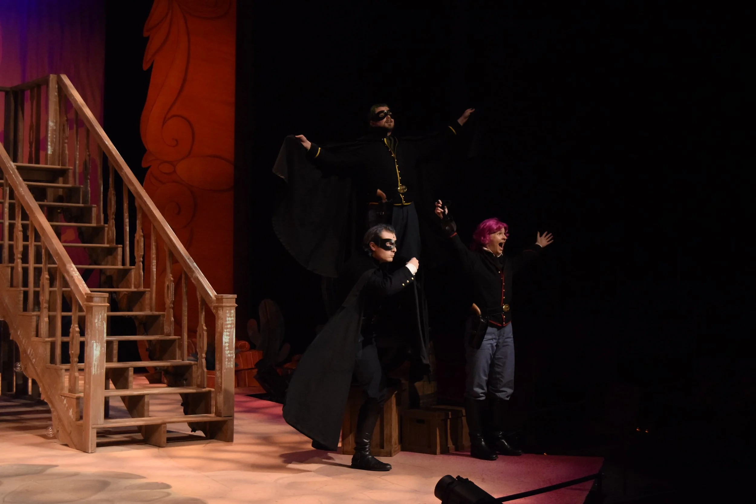 People dressed as characters from a theatrical performance on stage, with wooden stairs and colorful backdrop, some wearing masks and costumes.
