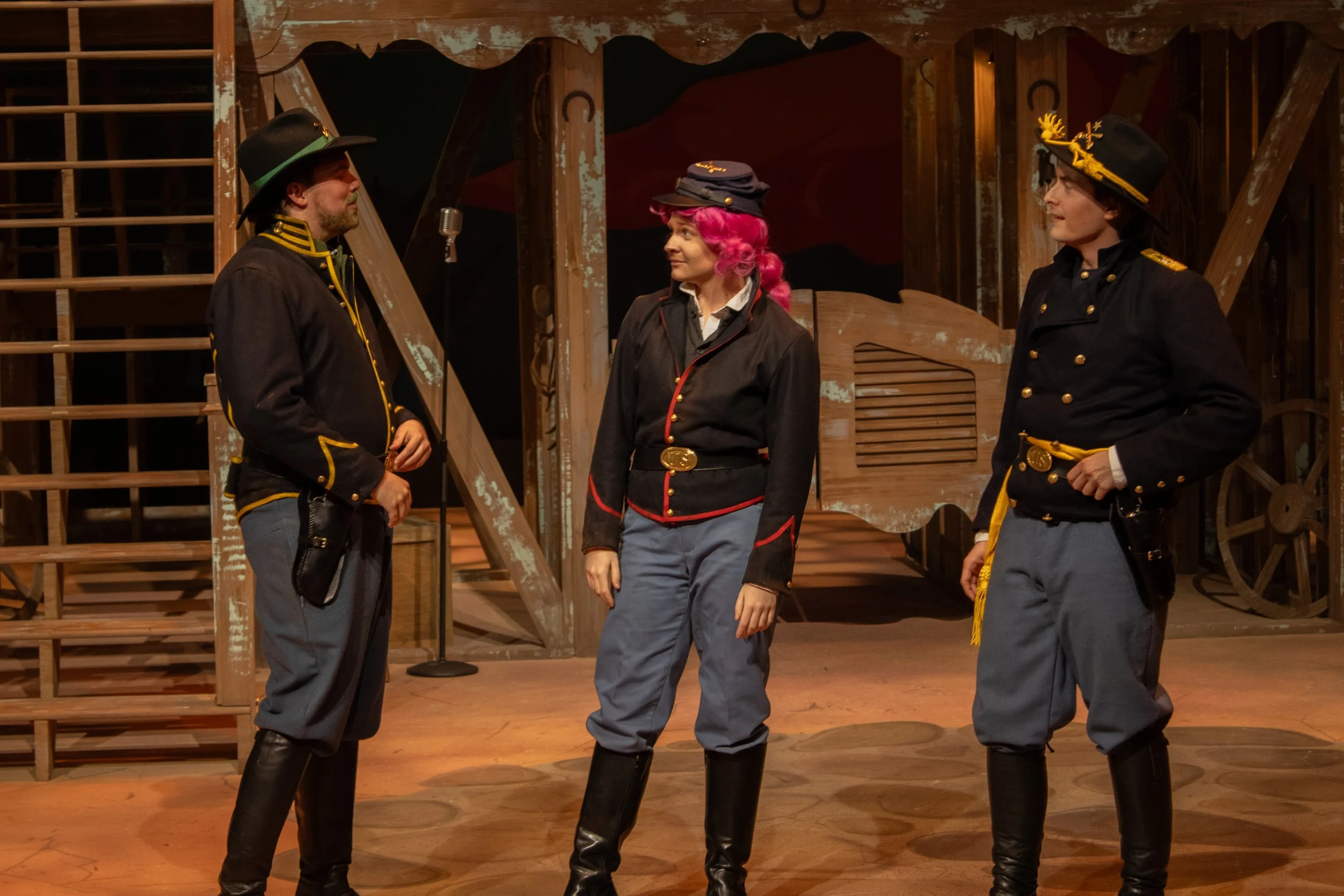 Three actors on stage in military-style costumes, engaged in a scene, with wooden props and a painted backdrop.