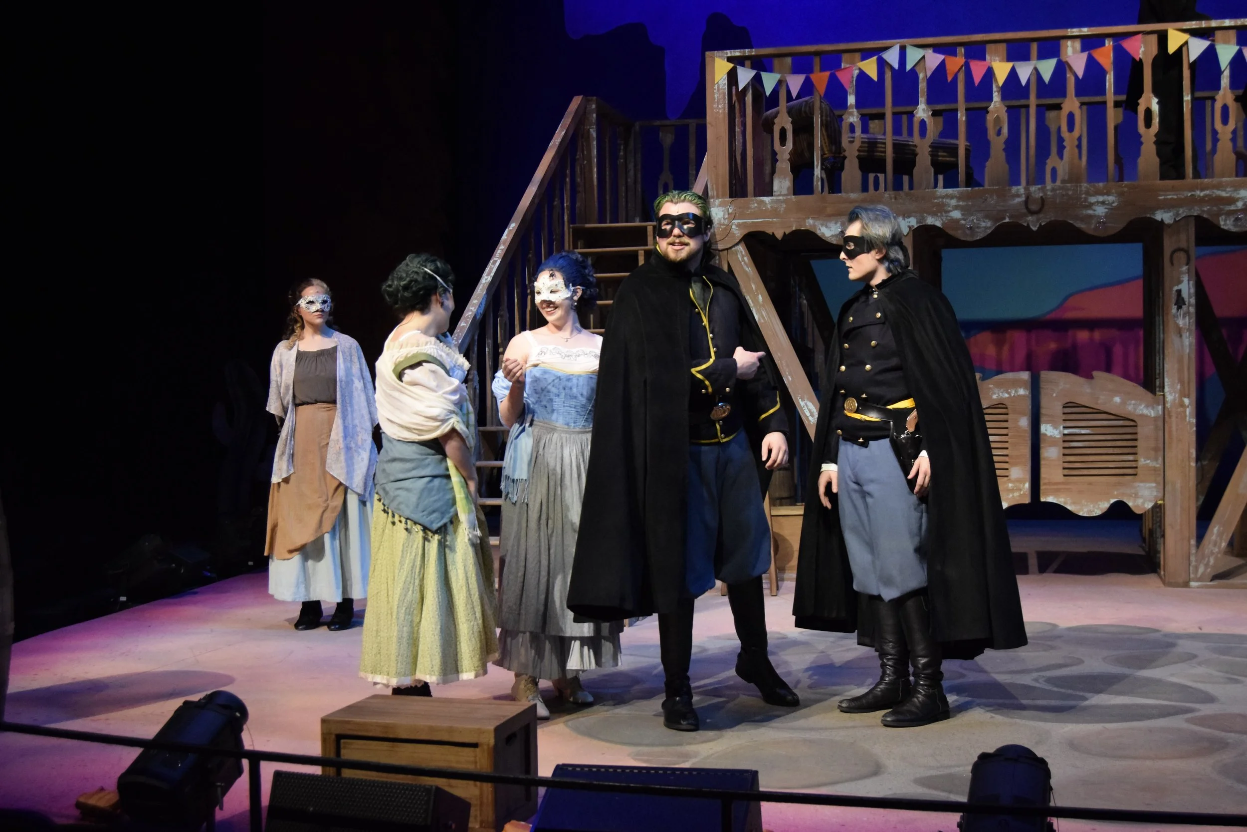 Actors dressed as characters in a theatrical performance, with three wearing masks, standing on stage near a wooden staircase decorated with colorful bunting in a theater setting.