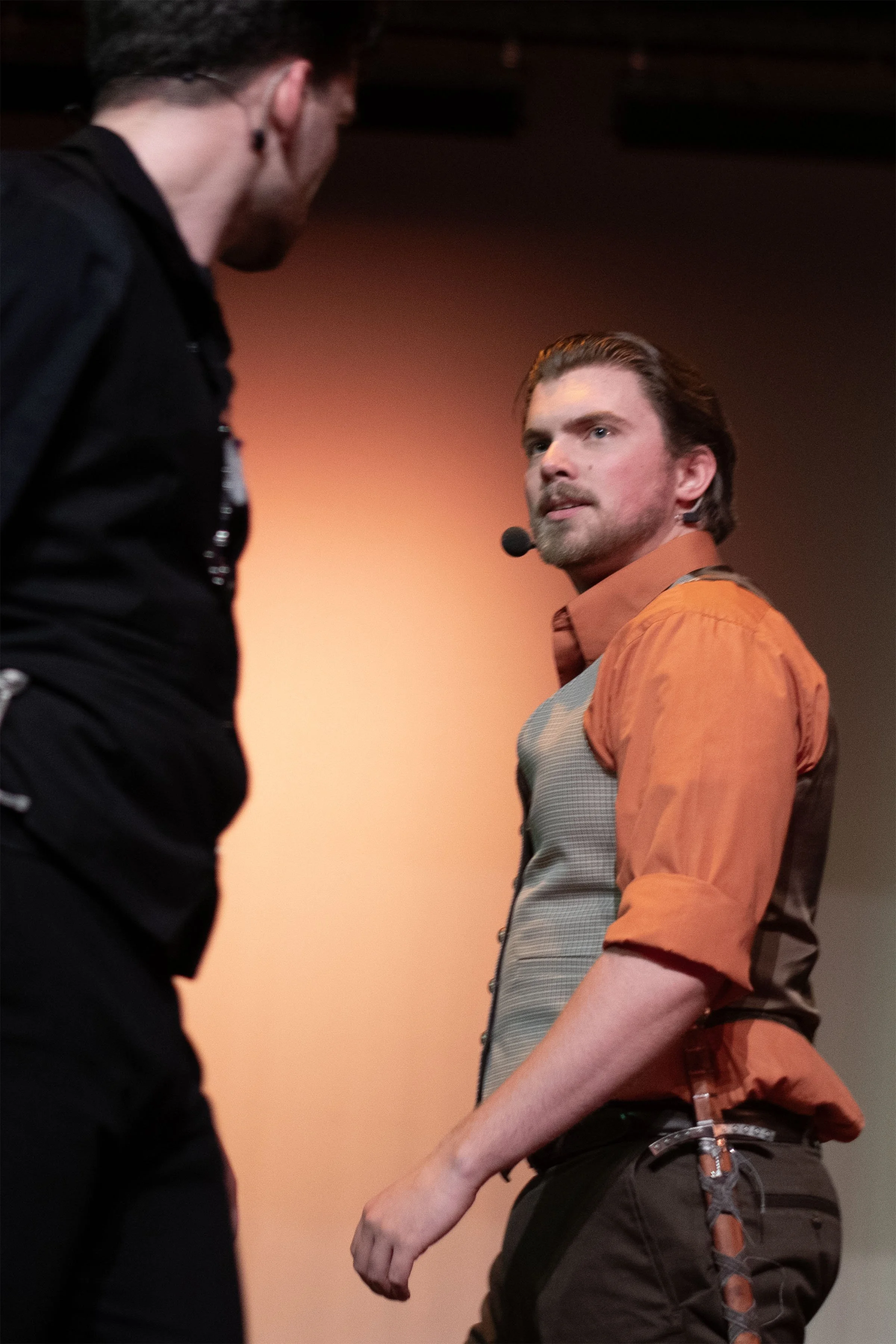 A man with a beard and styled hair wearing an orange shirt with rolled-up sleeves, a patterned gray vest, and brown pants, looking serious on stage with a microphone headset. Another person, partially visible, faces him.