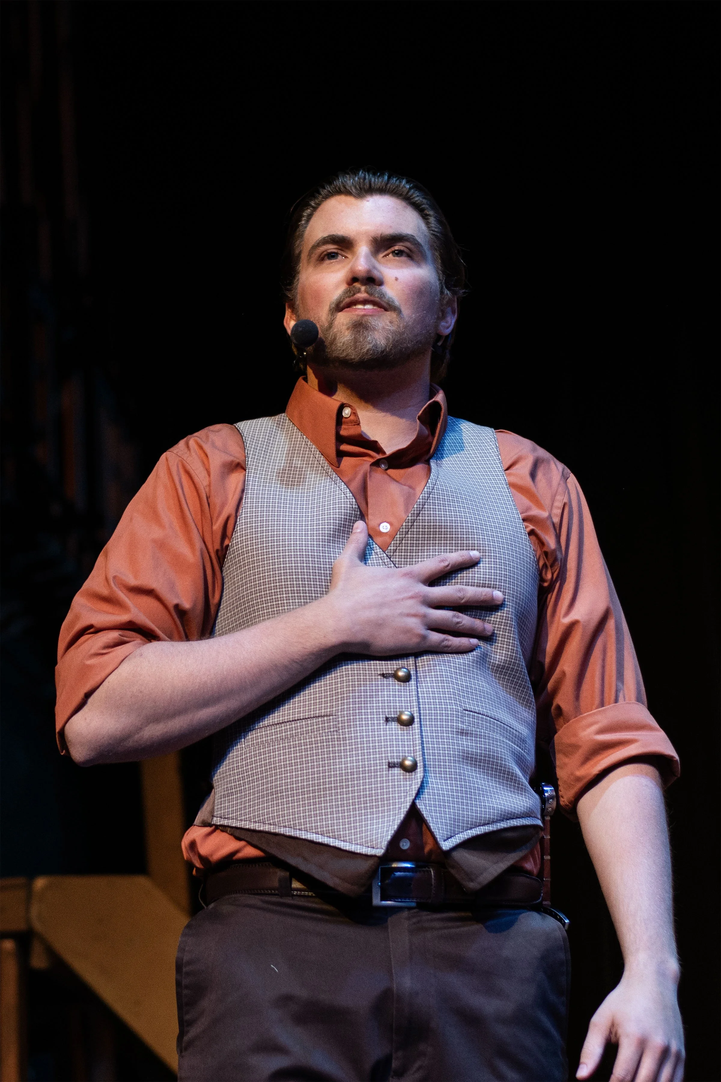Man on stage with hand over chest, wearing orange shirt, gray vest, and dark pants, speaking into a headset microphone.