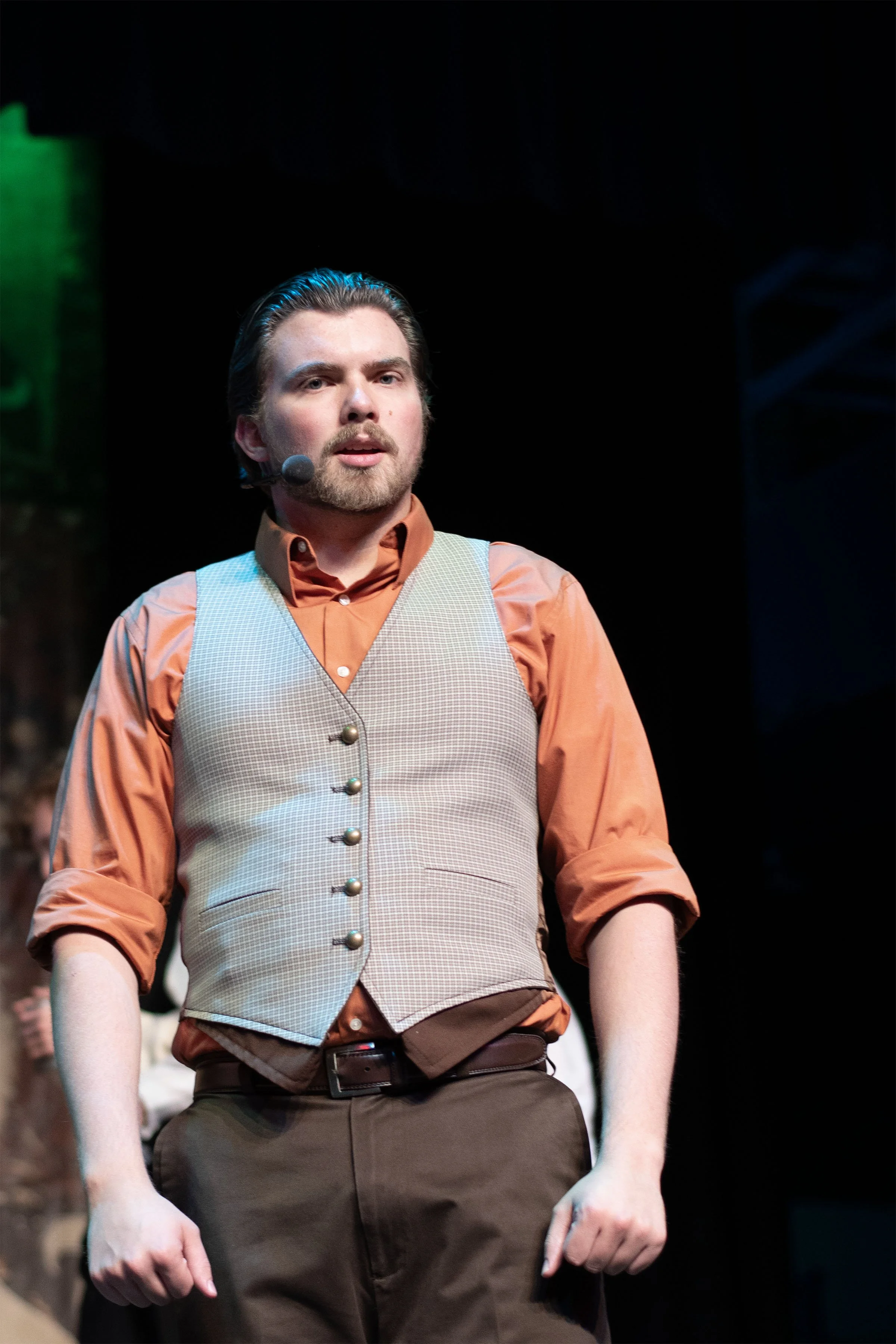 A man standing on stage, wearing a brown vest, orange shirt, and brown pants, with a microphone headset, during a theatrical performance.