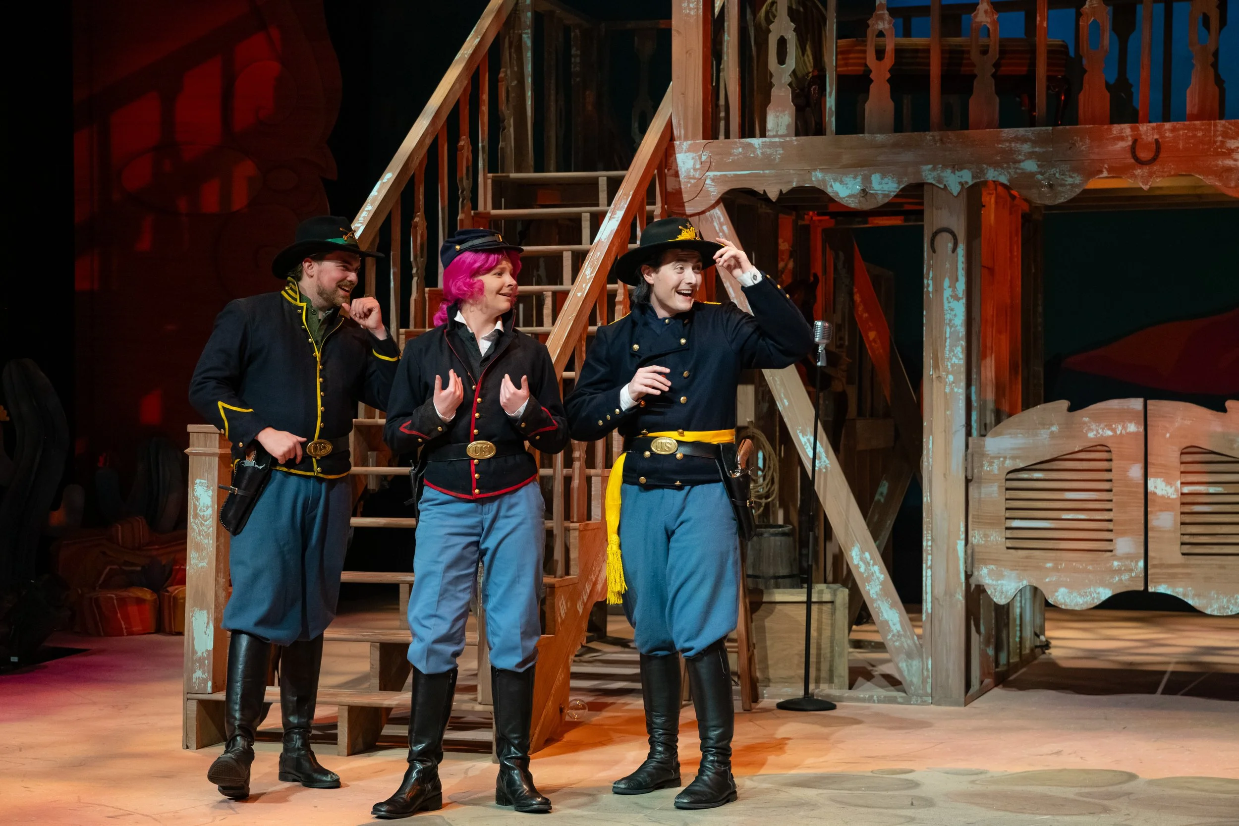 Three actors wearing cowboy costumes with hats and boots on stage, smiling and interacting during a theatrical performance, with a wooden set resembling a cabin or farm.