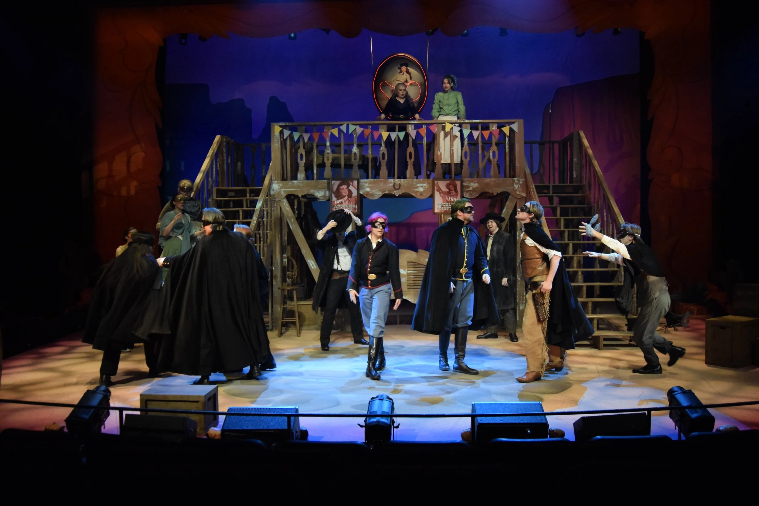 Theatrical stage scene with actors dressed as bandits and cowboys, on a rustic wooden set with stairs, bunting, and stage lights, during a theatrical performance.