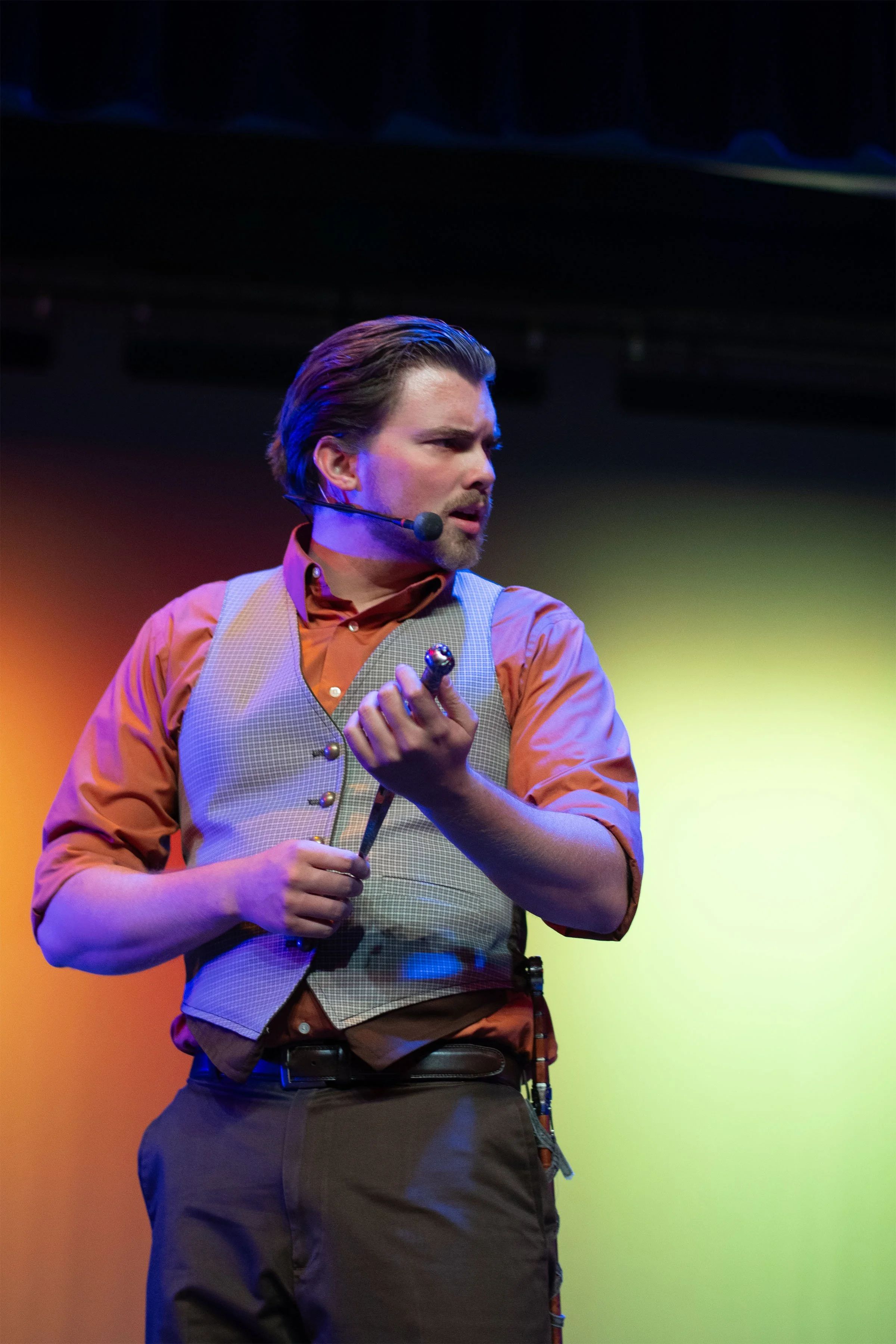 A man with a headset microphone, wearing a checkered vest over a brown shirt, holding an object in his right hand, and looking to his left on stage with colorful background lighting.