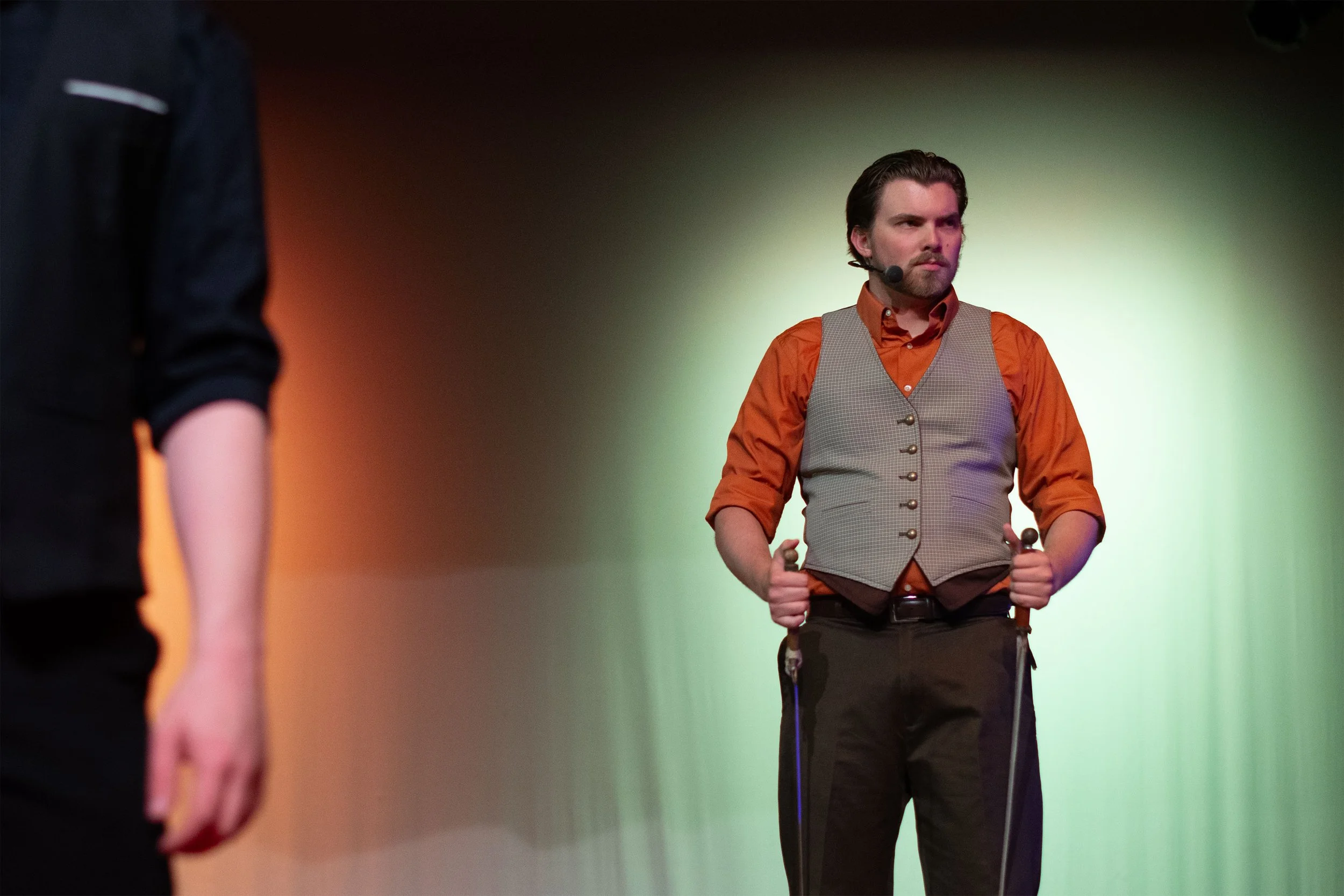 A man with a beard wearing an orange shirt and a gray vest stands with crutches on a stage, looking serious. A speaker is visible on his right, and the background is lit with green and orange lighting.