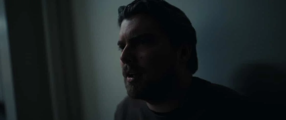 A close-up of a man with dark hair and a beard, looking out a window in a dimly lit room.