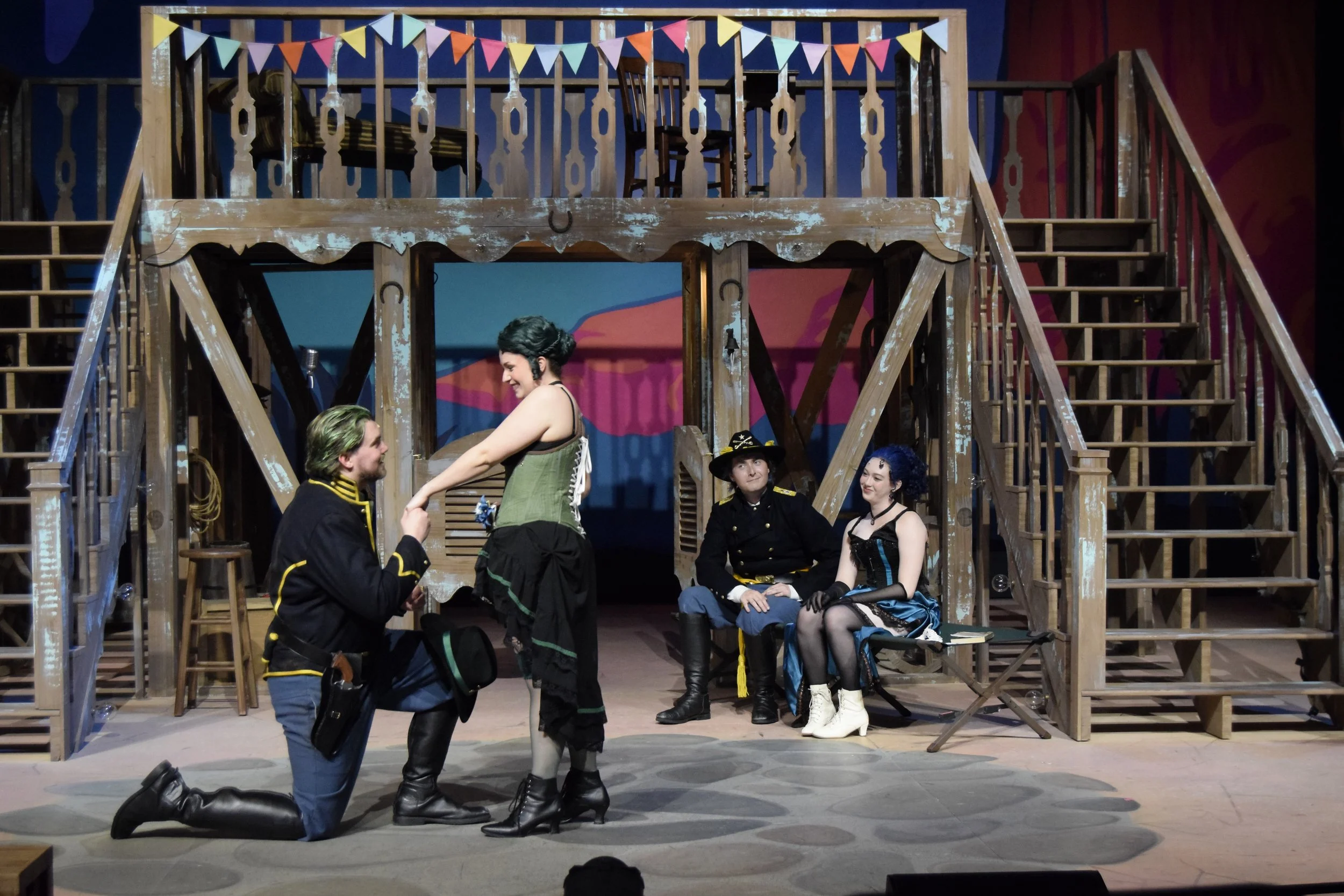 The image shows a theatrical stage with actors in costumes, suggesting a scene from a play or musical. Some actors are seated, and others are engaged in a scene involving kneeling and standing positions. The stage set includes a rustic wooden structu