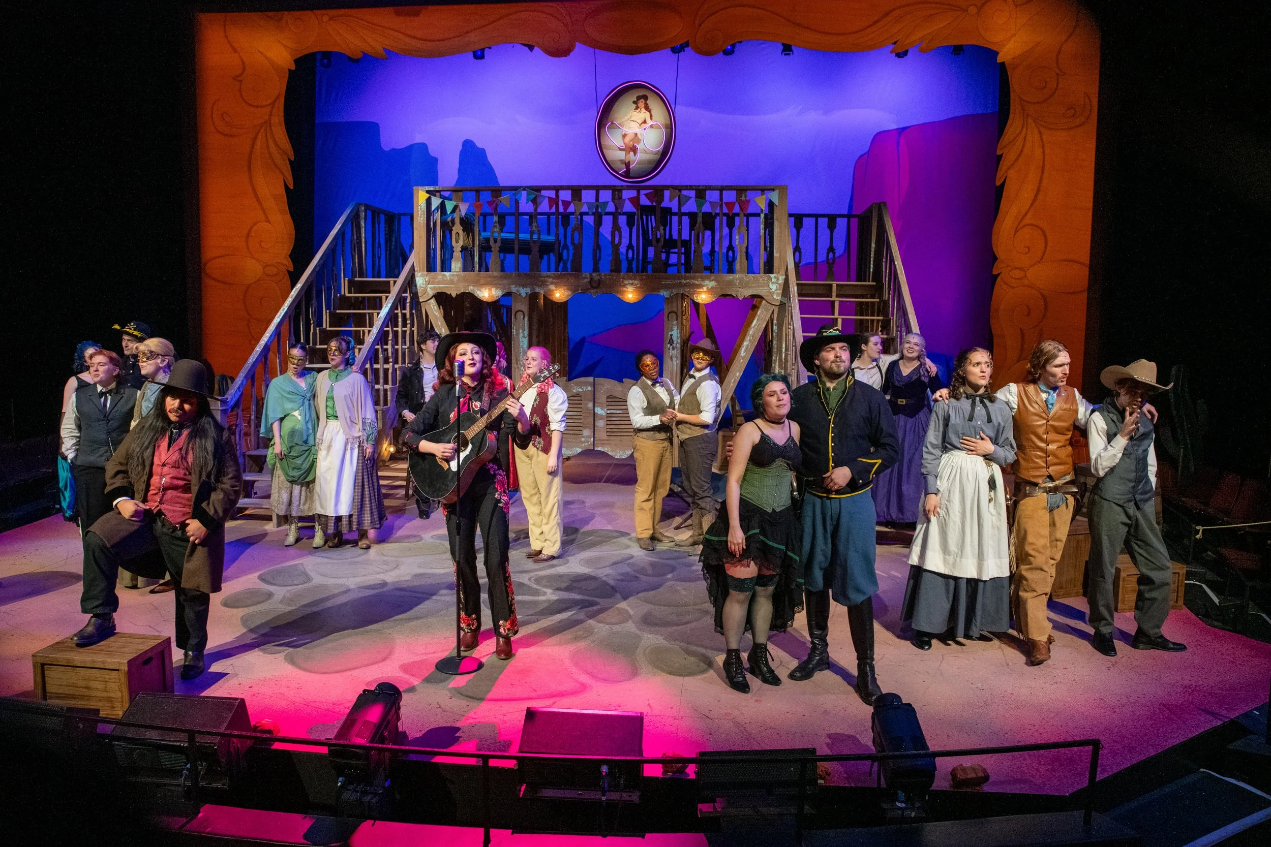 A group of performers on stage in a theater, with some dressed in Western costumes and others in vintage or theatrical attire, standing in front of a colorful set with a raised platform, stairs, and a painted backdrop depicting a landscape.