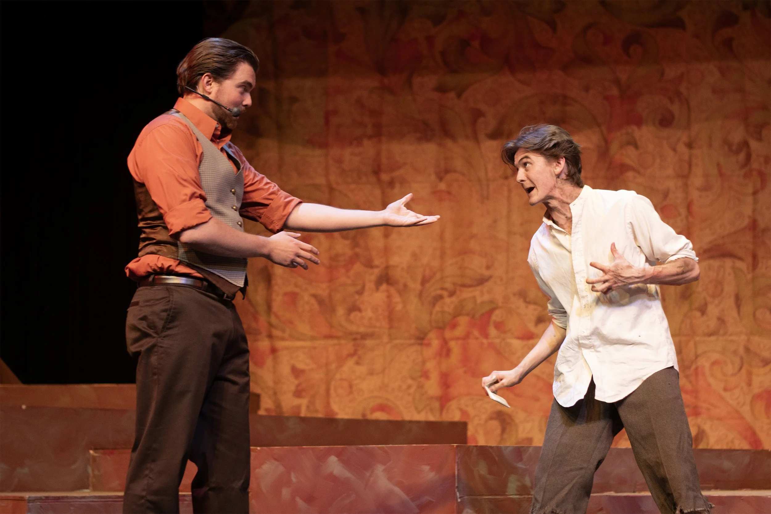 Two actors performing on stage, expressing emotion. The actor on the left is wearing an orange shirt, brown pants, and a gray vest, extending his hand. The actor on the right is wearing a white shirt and brown pants, and appears to be speaking passionately.