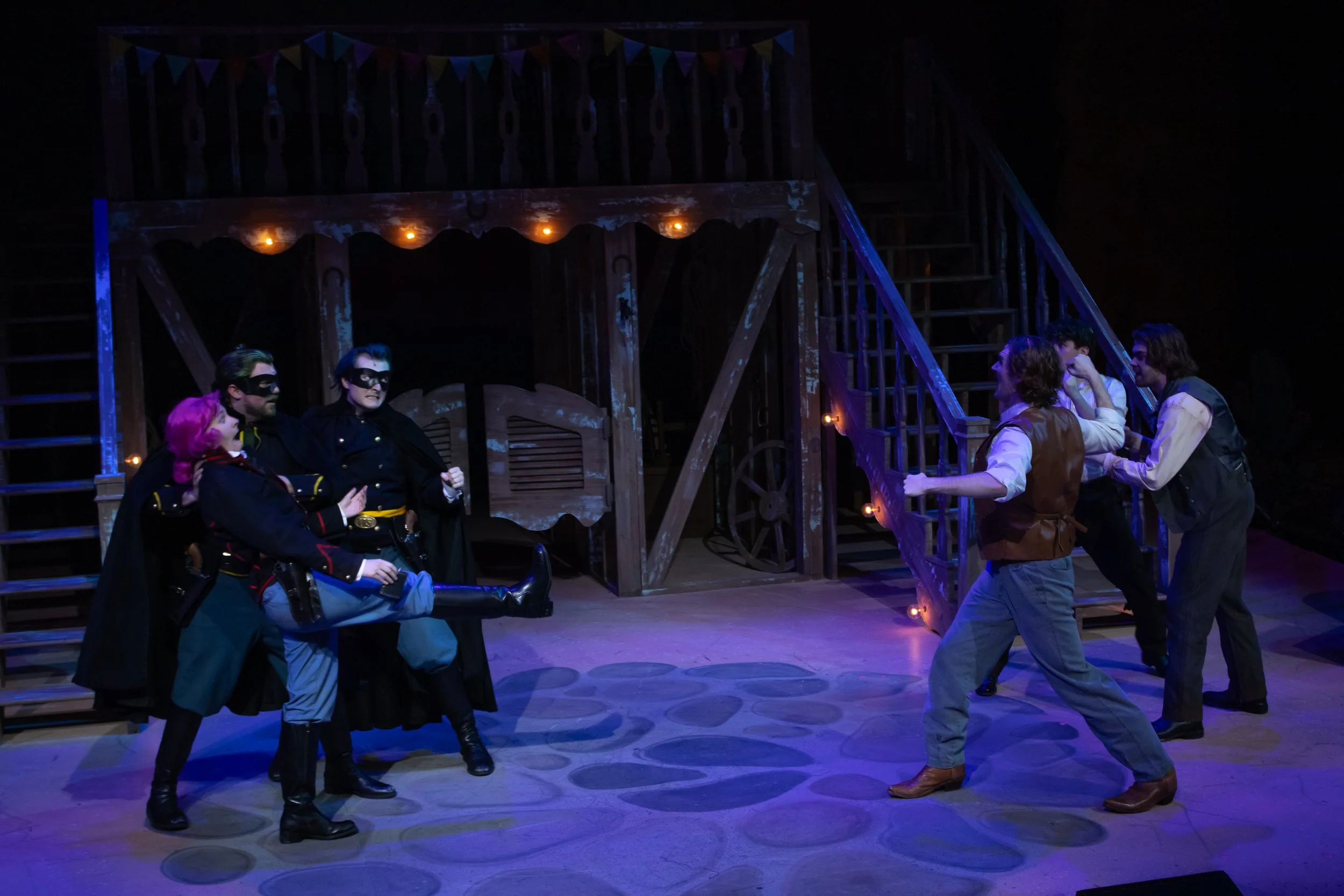 The image shows a stage scene from a theatrical production resembling the play 'The Three Musketeers.' There are six actors visible, four dressed as musketeers with swords, and two others engaged in a conversation. The stage has a rustic wooden backdrop with stairs and bunting lights strung overhead.