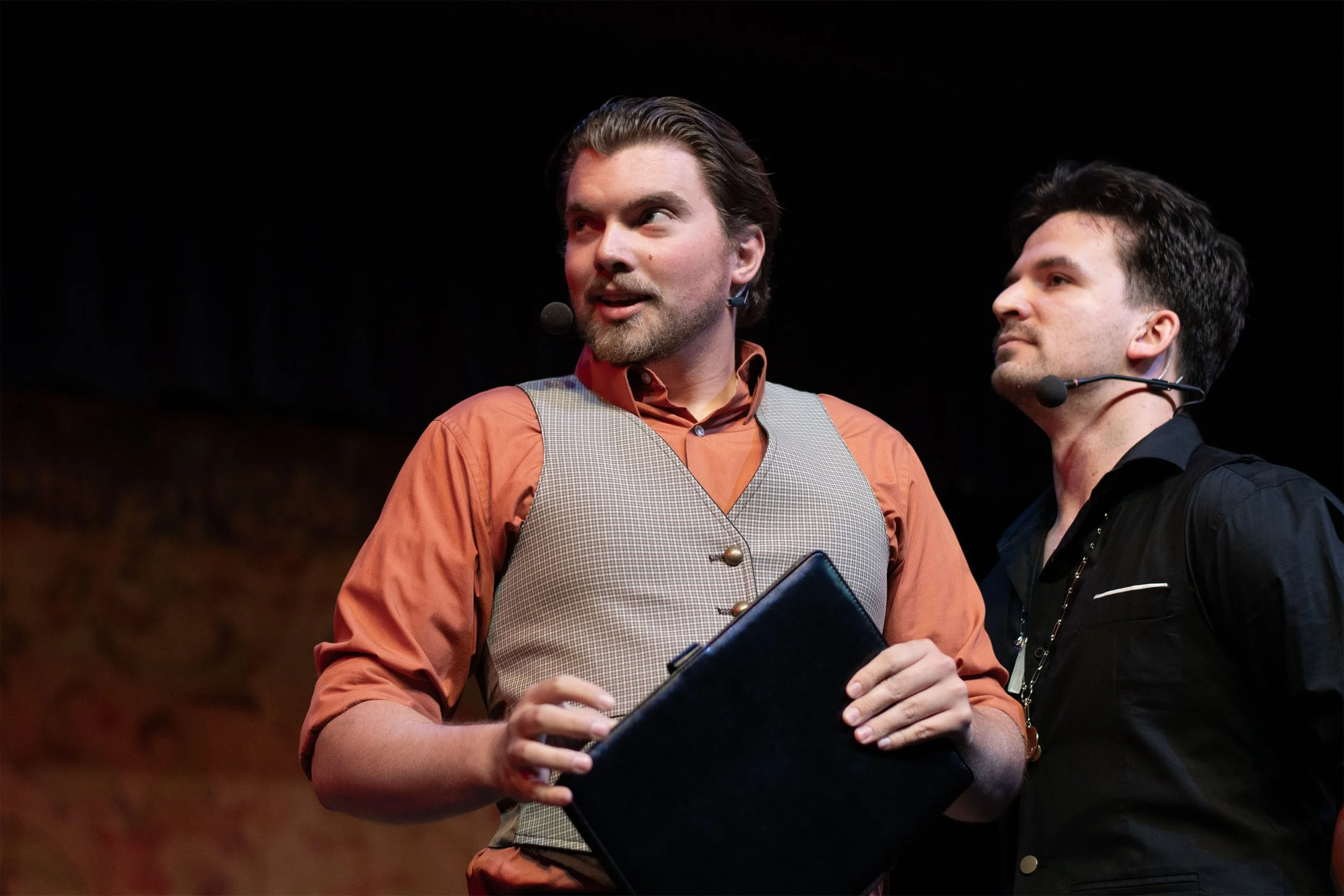 Two men standing on stage during a performance, both wearing microphones. The man on the left is holding a black folder, wearing a rust-colored shirt with a gray vest, and the man on the right is in a black shirt with glasses.
