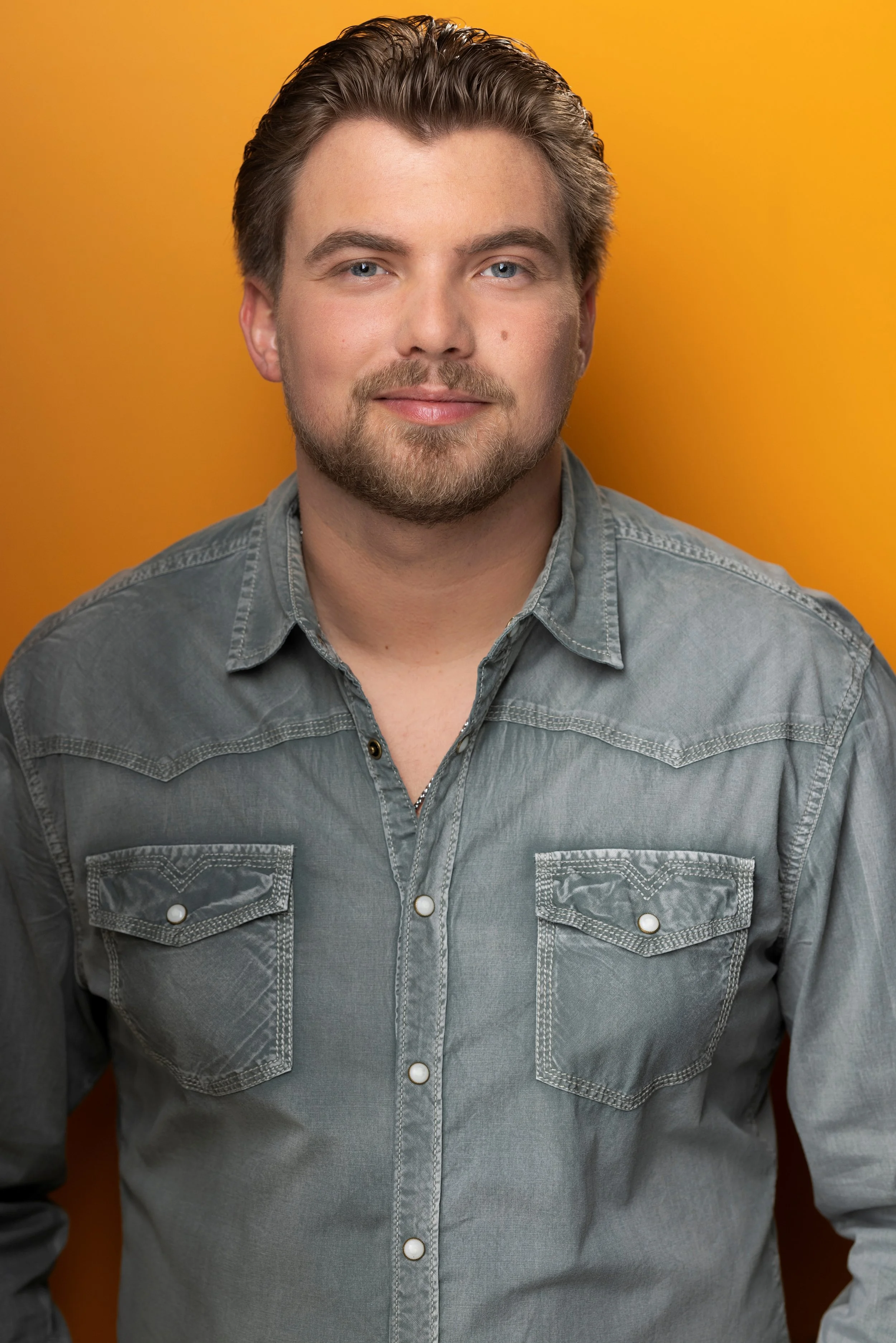A man with light skin, blue eyes, and dark blonde hair styled back, wearing a gray denim button-up shirt, standing against a yellow background.