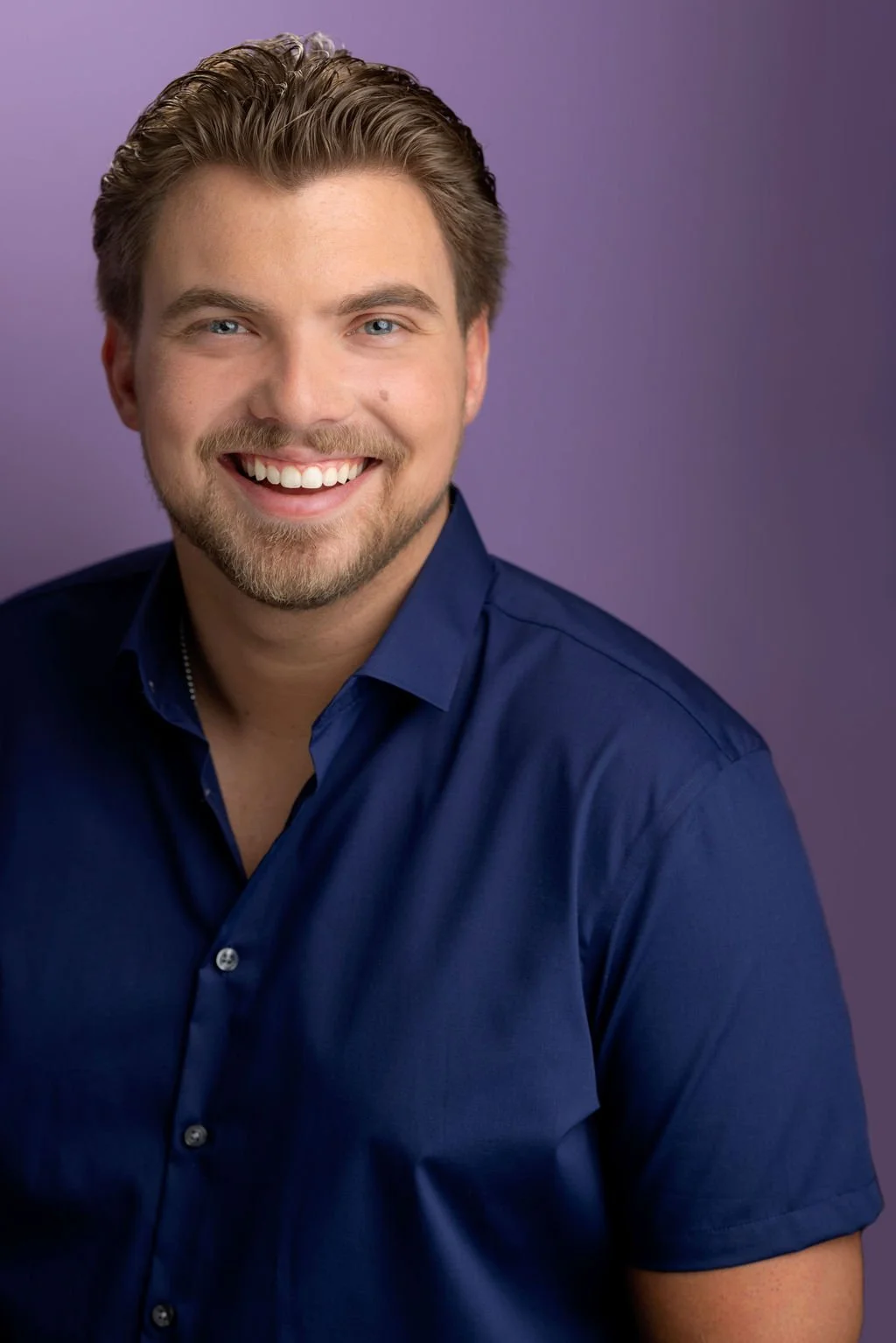 A smiling young man with light brown hair, blue eyes, and a beard and mustache, wearing a dark blue button-up shirt, against a purple background.