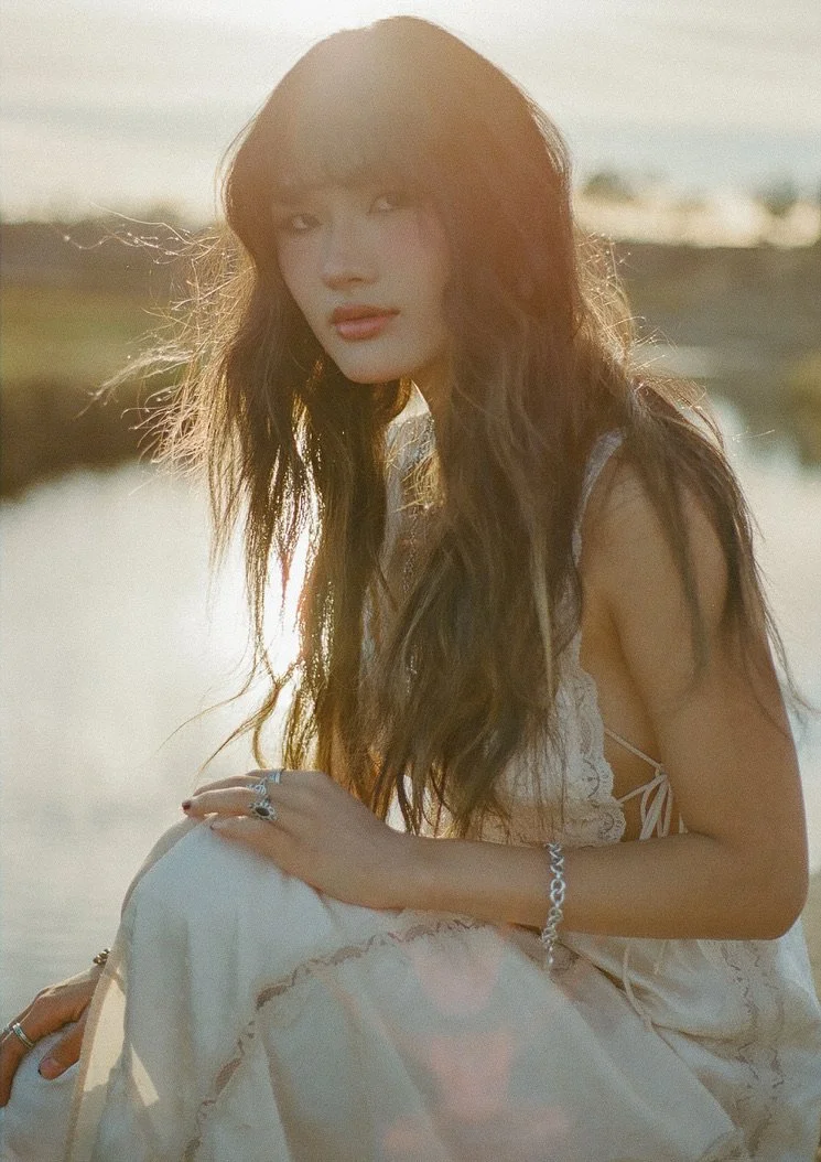 A woman with long brown hair, wearing a light-colored dress, sitting outdoors near the water during sunset.