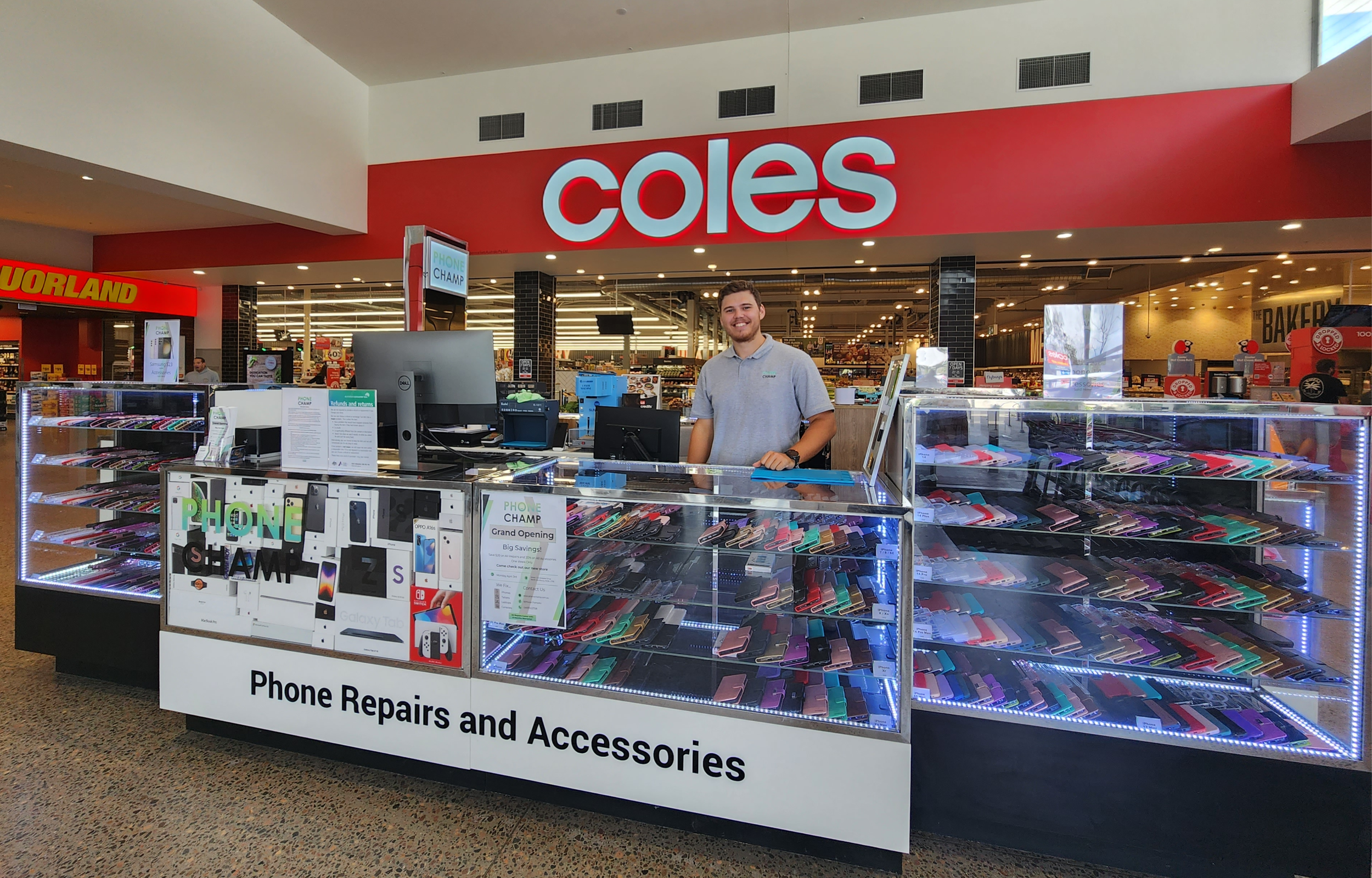 Inside a Coles supermarket, a mobile phone accessories kiosk with a man smiling behind the counter surrounded by colorful phone cases and accessories.