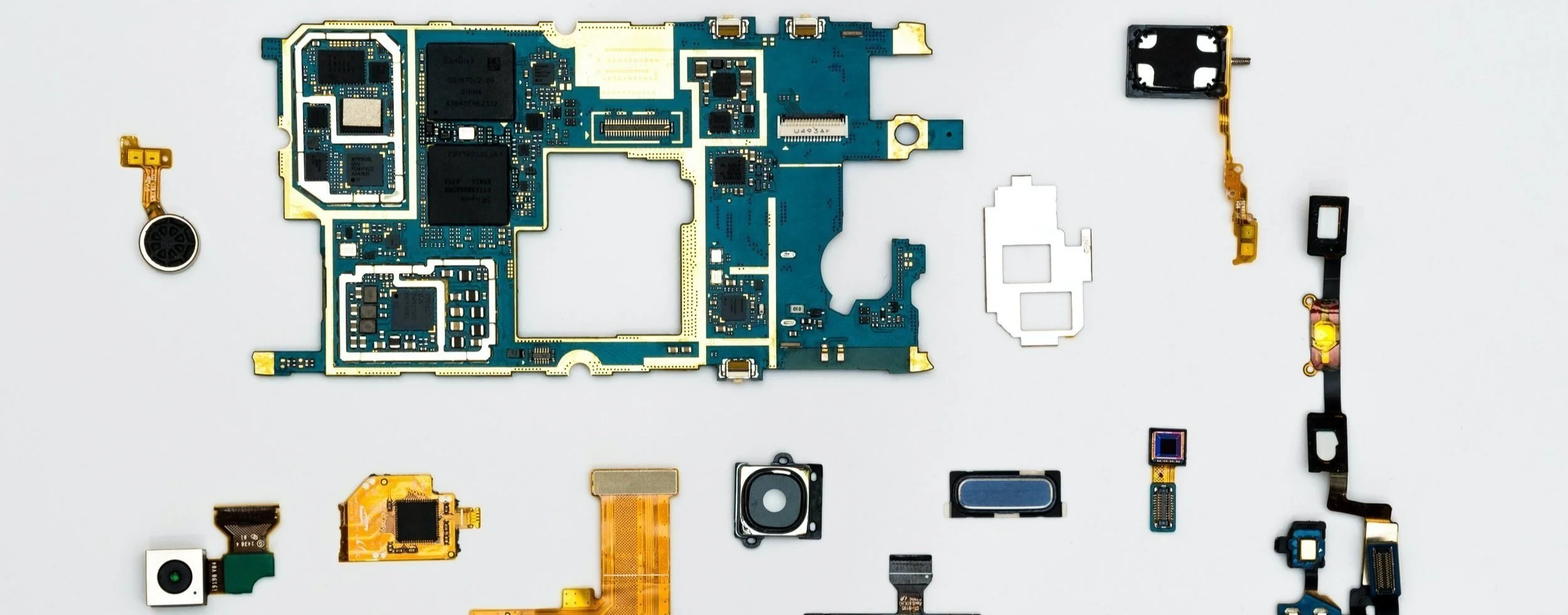 Disassembled electronic circuit board components including a large blue main board, smaller circuit parts, flex cables, and connectors arranged on a white background.