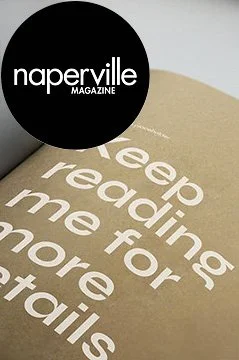 Close-up of the cover of a Naperville Magazine issue, with a black circle in the upper left corner and white text that reads "naperville MAGAZINE." The background shows part of a beige book or magazine cover with white text that says, "Keep reading me for more details."