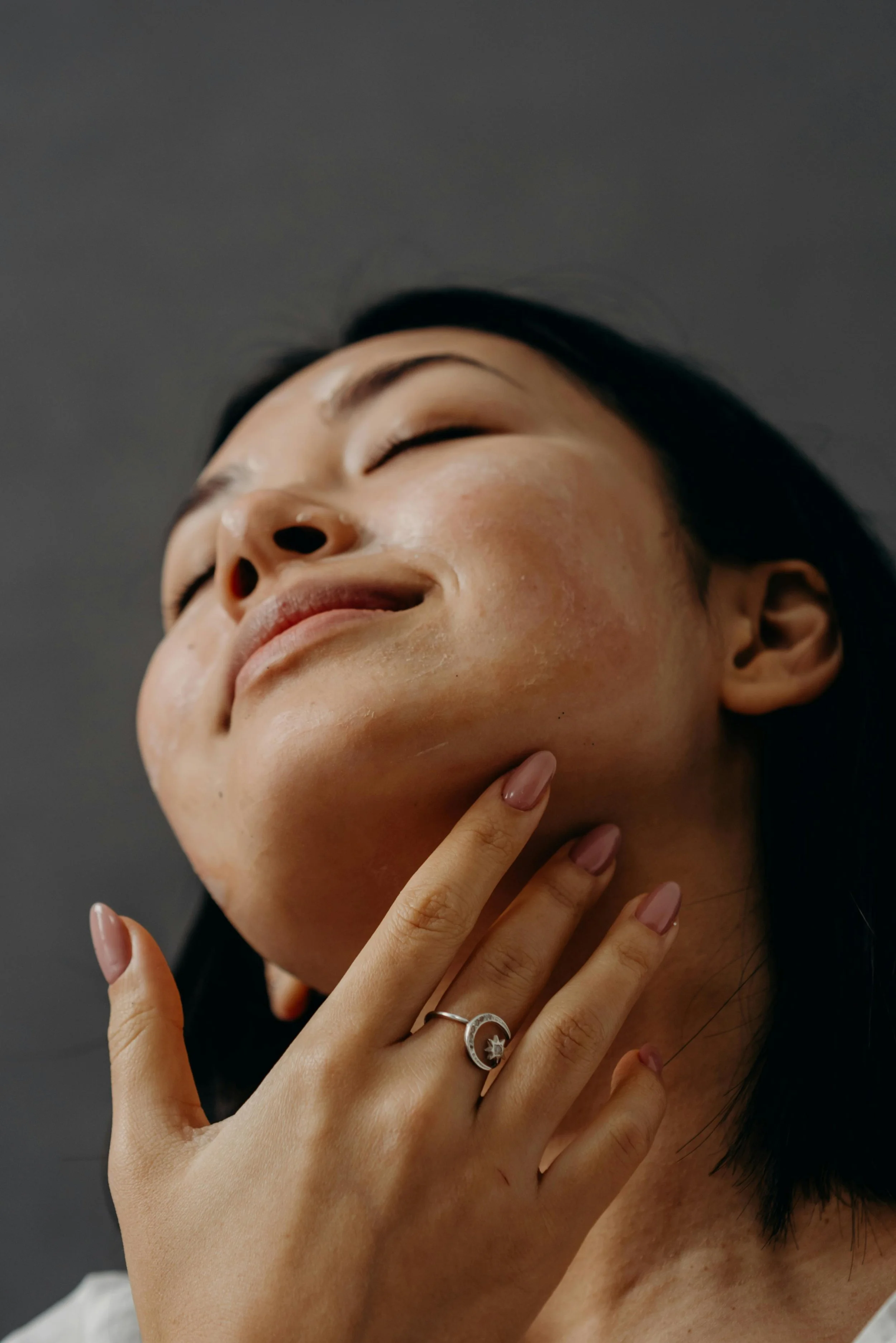 A woman with short dark hair is touching her chin with her hand, which has a silver ring with a moon and star design. She appears to be enjoying a skincare treatment, with her eyes closed and a serene expression on her face.