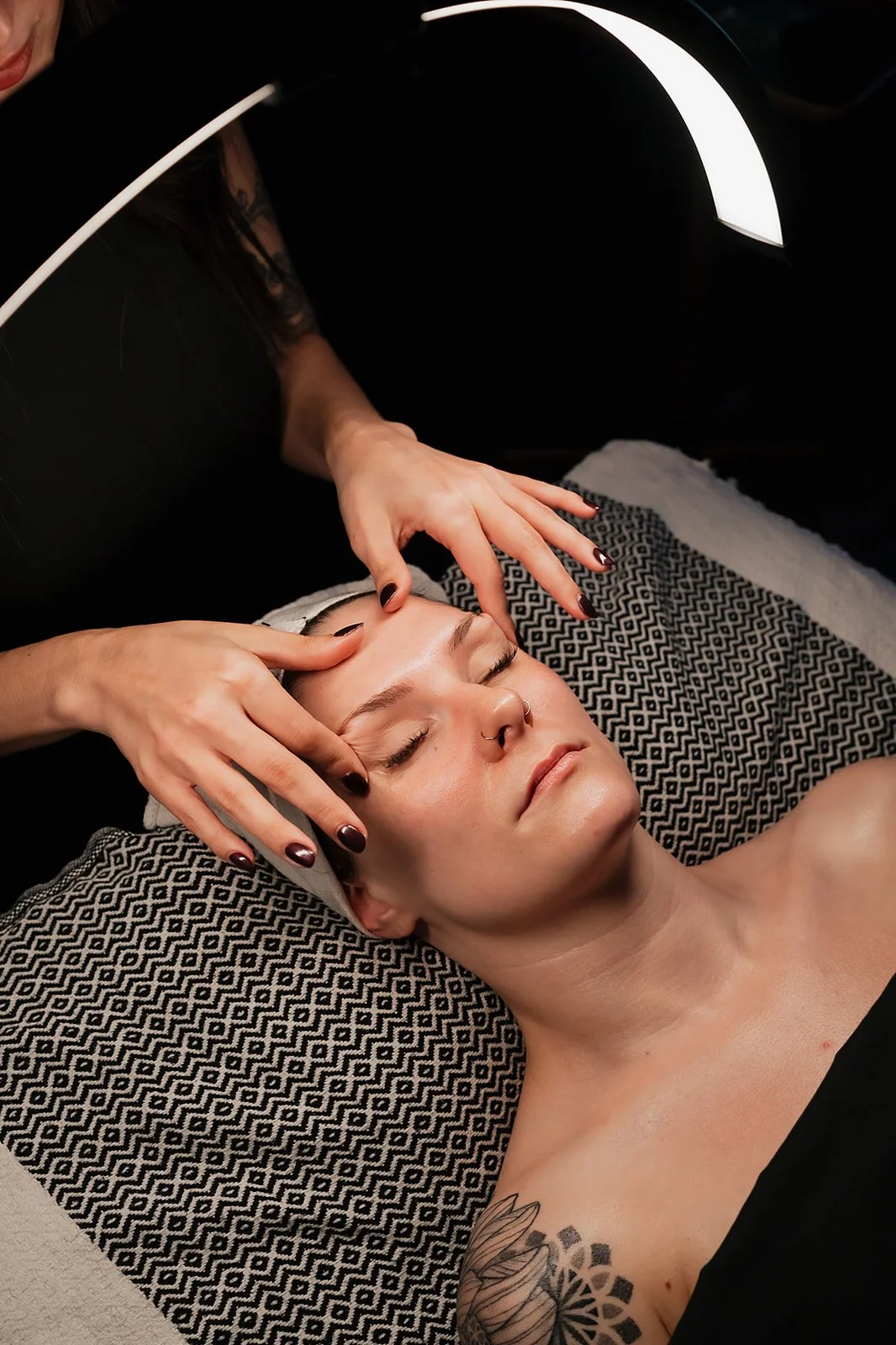A woman receiving a relaxing facial treatment lying on a table with her eyes closed, while Amanda Jerman licensed esthetician and owner of Healing Vibes Esthetics Denver uses their hands to massage her forehead and face.