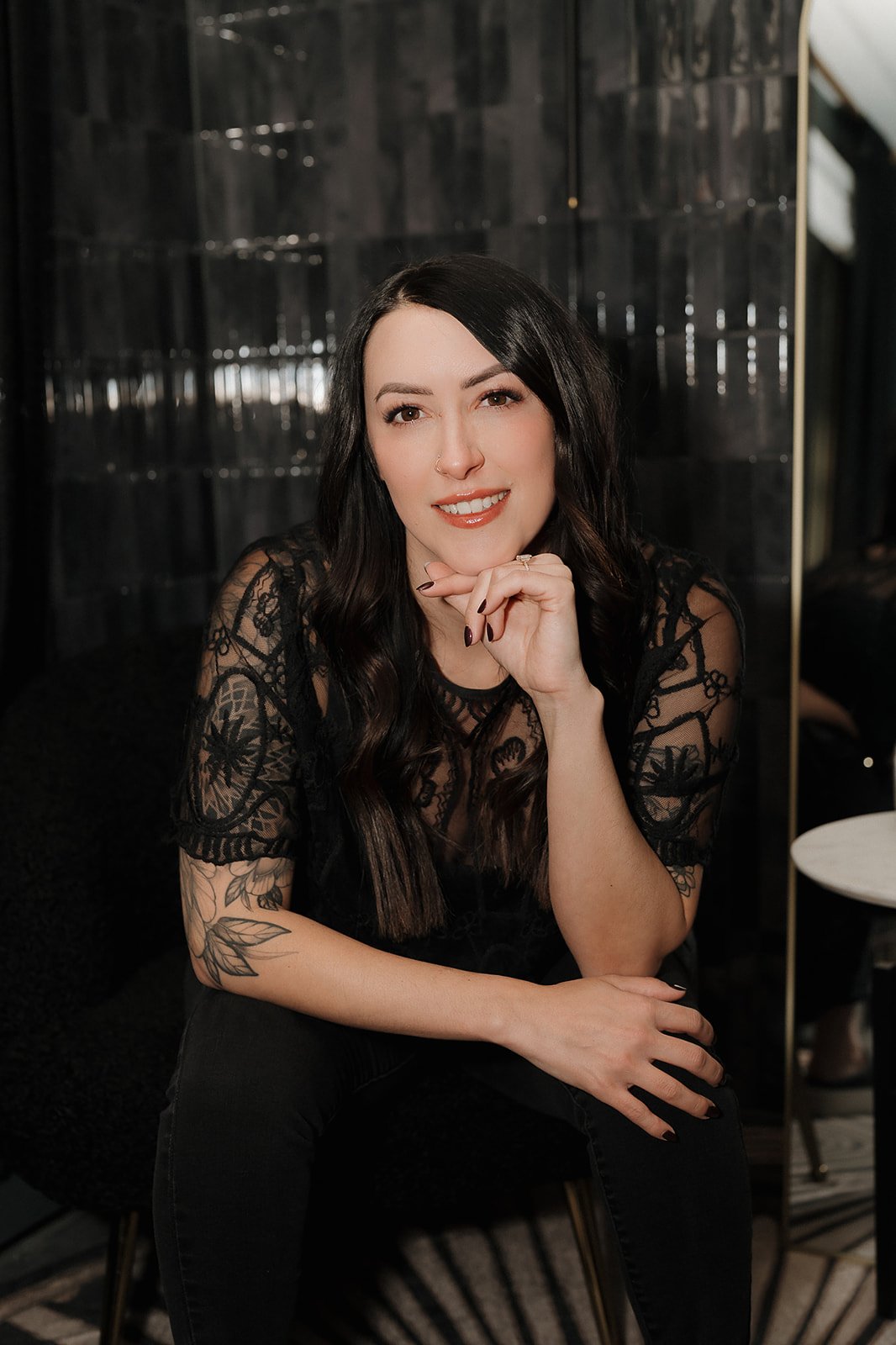 Amanda Jerman, licensed esthetician and owner of Healing Vibes Esthetics Denver wearing a black lace top, sitting on a chair with a dark background, smiling at the camera with one hand under her chin.