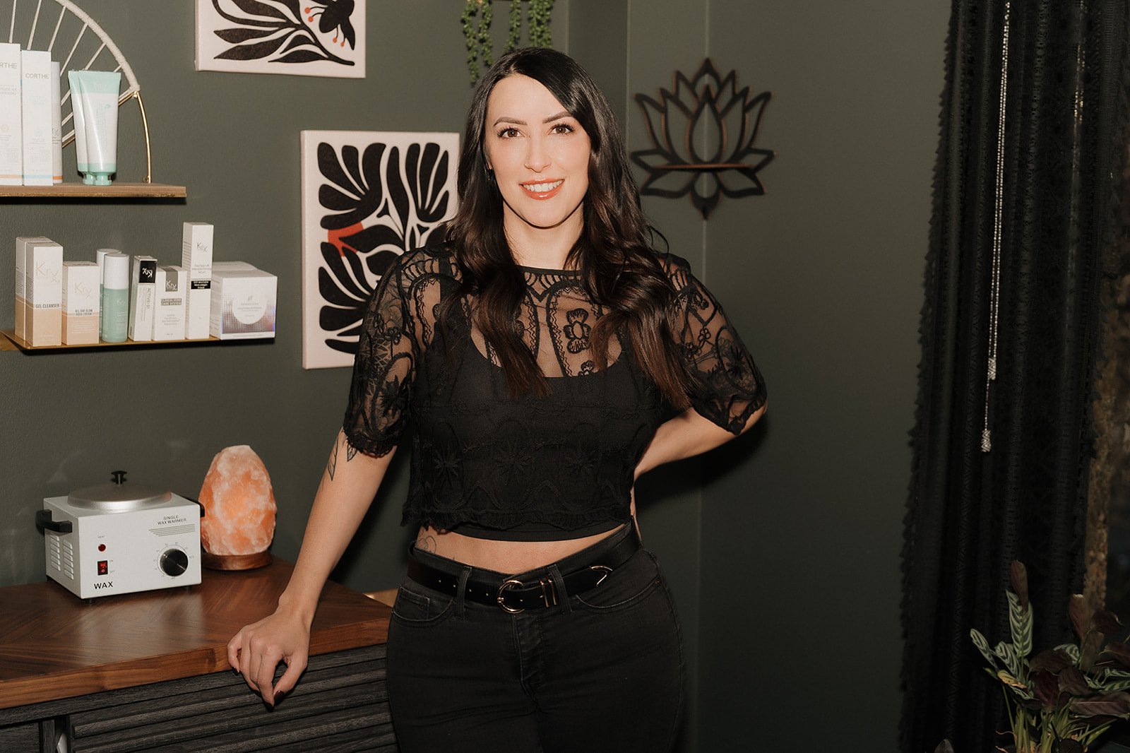 Amanda Jerman, licensed esthetician and owner of Healing Vibes Esthetics wearing a black lace crop top and black pants, standing in a room with dark green walls decorated with black and white artwork, a salt lamp, and shelves with skincare products.
