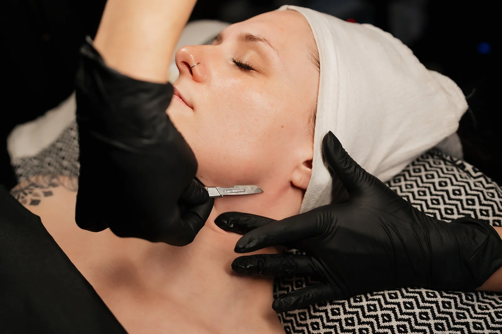 A woman receiving a dermaplane facial while lying down, with her eyes closed and wearing a white head covering. Amanda Jerman, licensed esthetician and owner of Healing Vibes Esthetics Denver wearing black gloves is working on her skin.