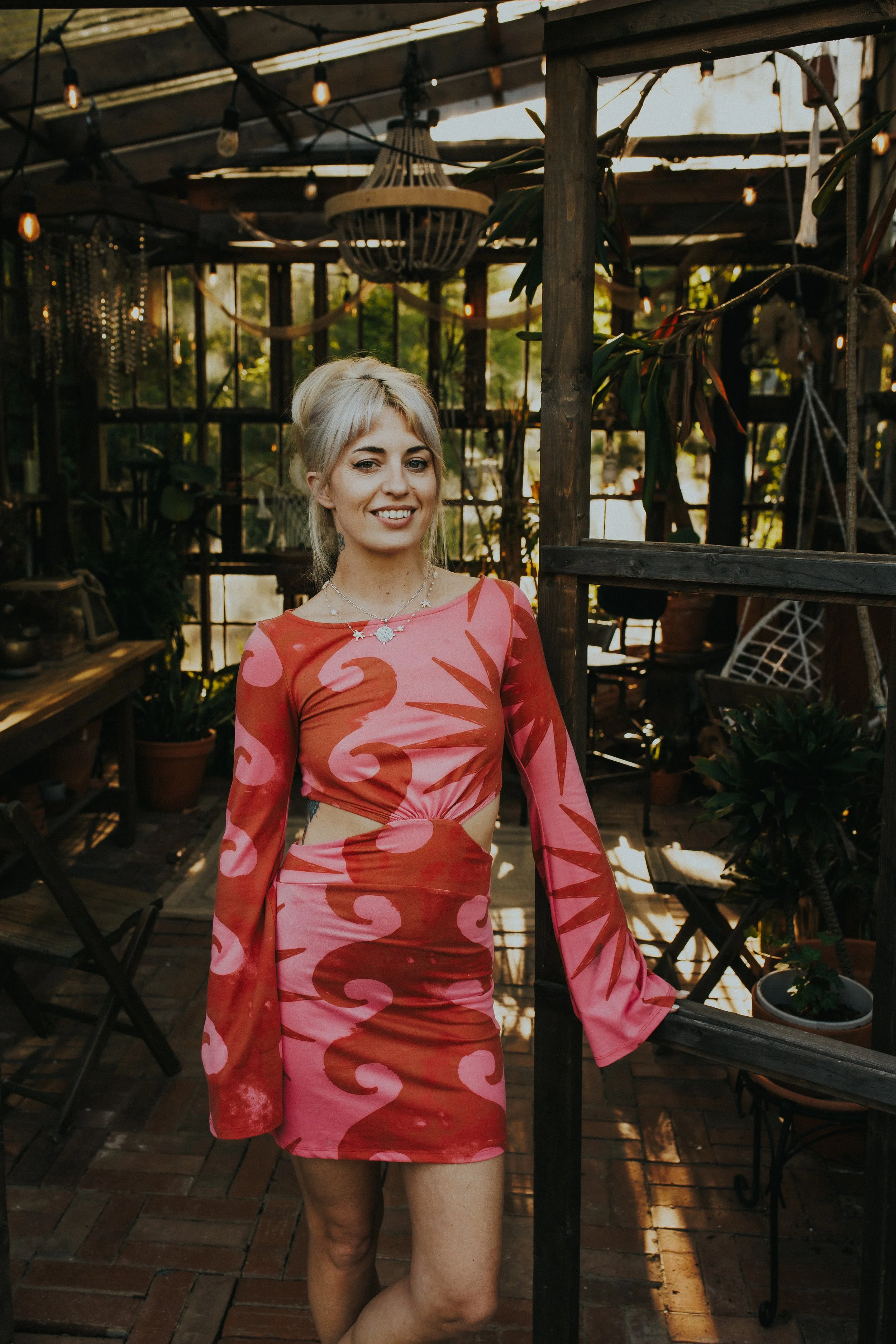 A woman with platinum blonde hair wearing a pink and red swirl pattern dress standing inside a wooden greenhouse with plants and decorative lights.