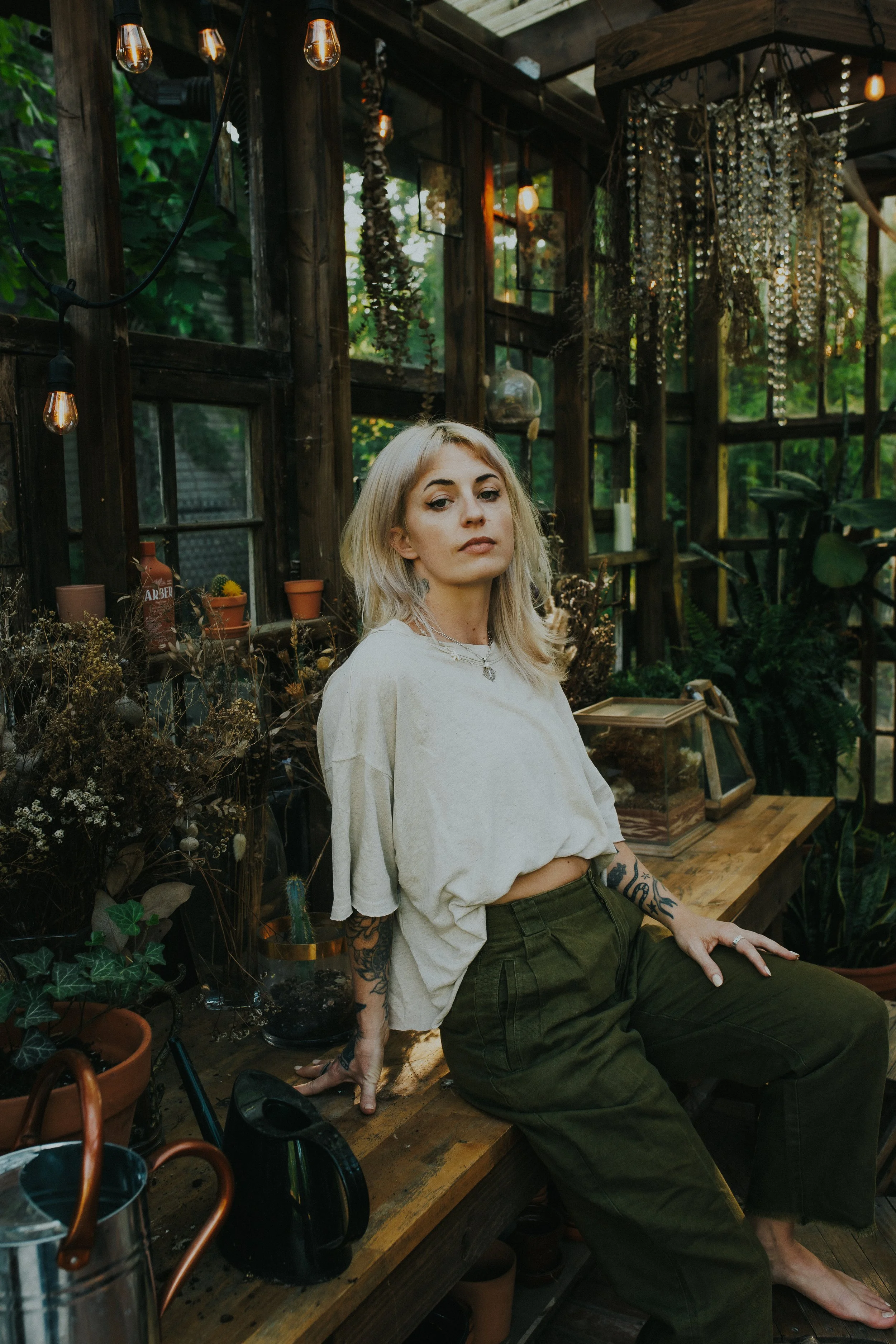 A woman in repose sitting on a wooden bench in a lush greenhouse with various potted plants and hanging decorations around her.
