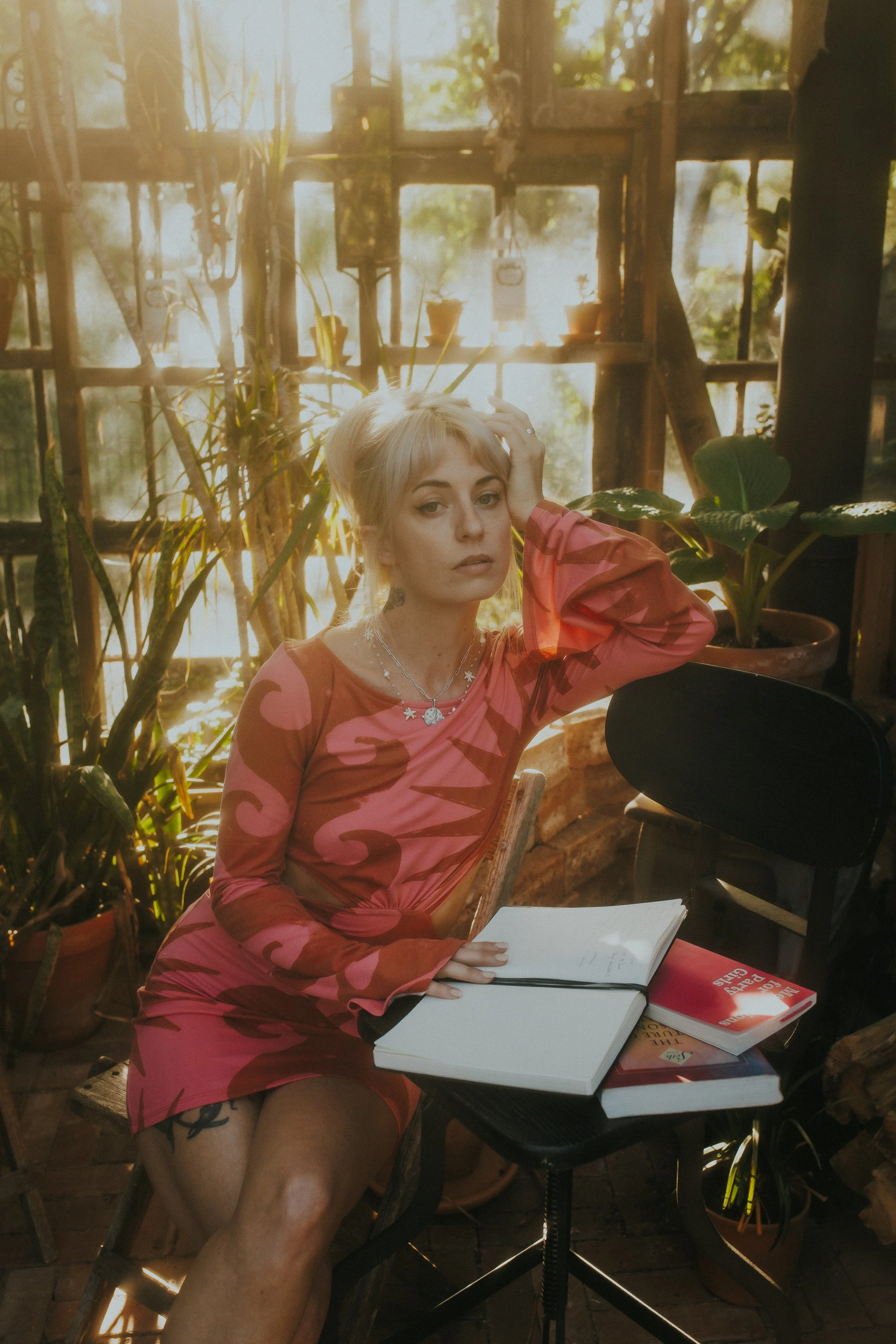 A woman sitting at a table in a cozy, rustic greenhouse filled with potted plants and sunlight streaming through large windows. She is wearing a pink dress with swirling patterns, has short blonde hair, and a contemplative expression while holding her hair with one hand. There are books and an open notebook on the table in front of her.